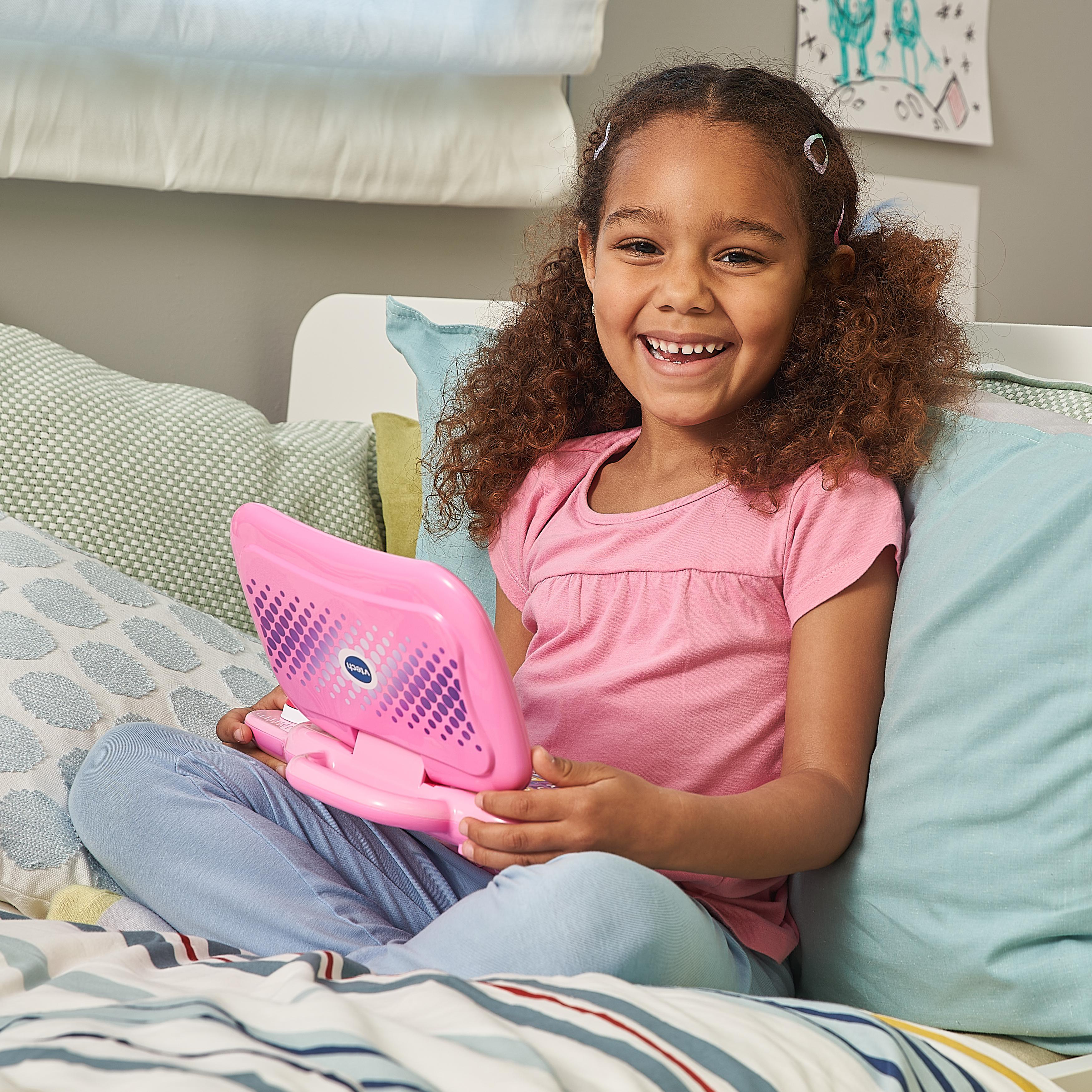 Mädchen mit lockigem Haar, das lächelt, hält einen rosa Spielzeuglaptop auf einem Bett mit Kissen.