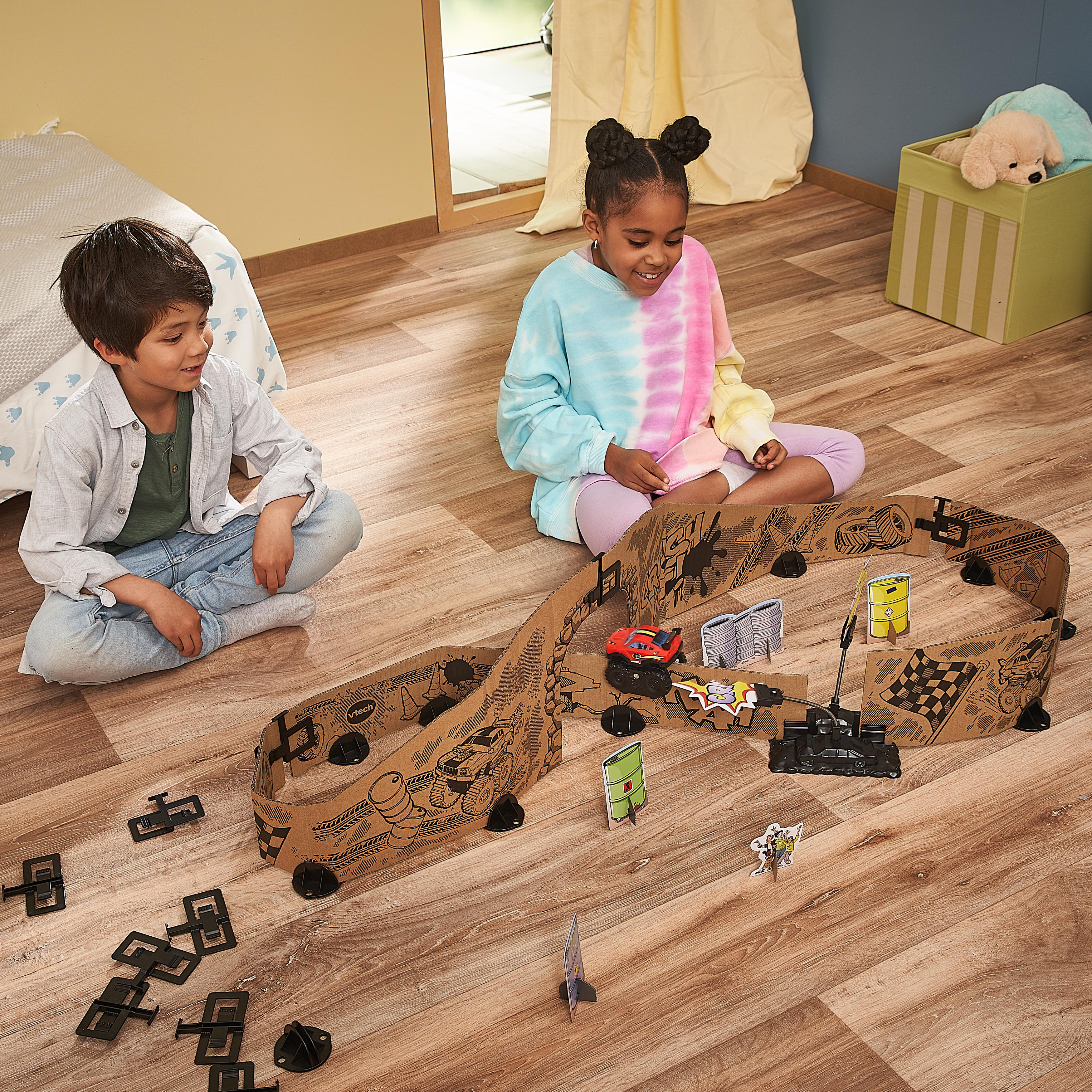 Zwei Kinder spielen mit Pappe-Rennstrecke, Autos und Hindernissen auf Holzboden.