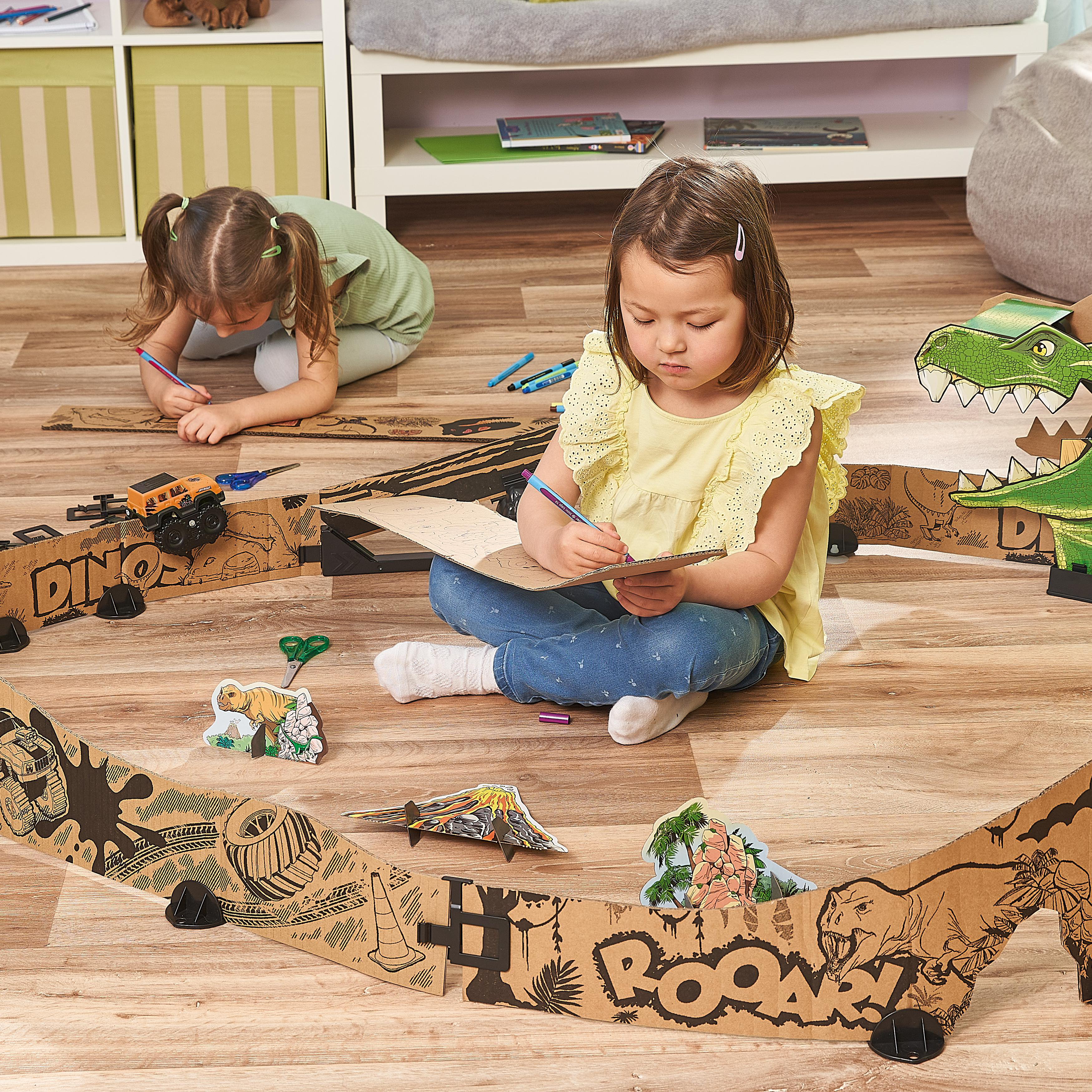 Zwei Kinder spielen mit einer Dinosaurier-Pappstrecke auf einem Holzboden.