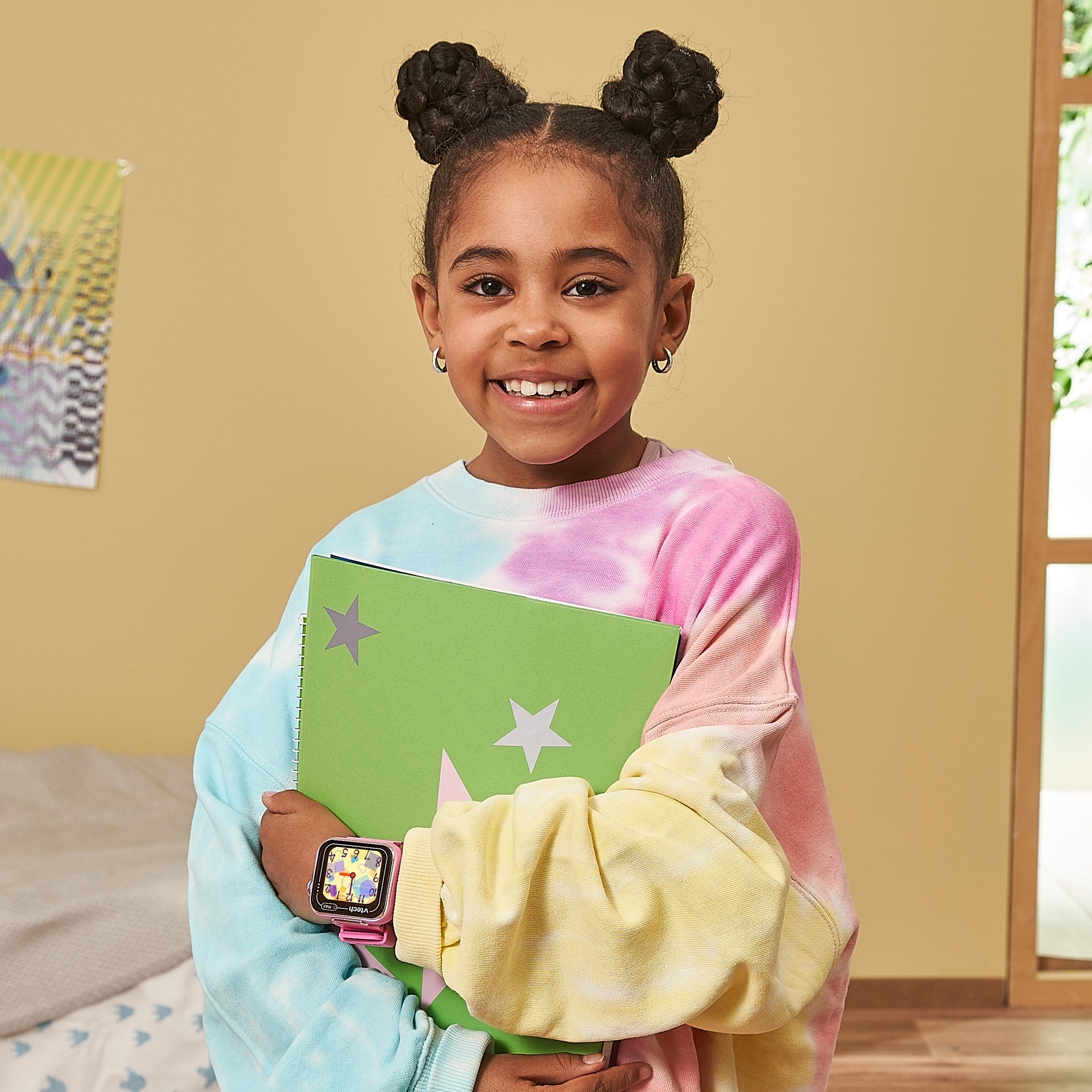 Ein lächelndes Mädchen mit Space Buns, das ein grünes Notizbuch hält, trägt einen bunten Pullover.