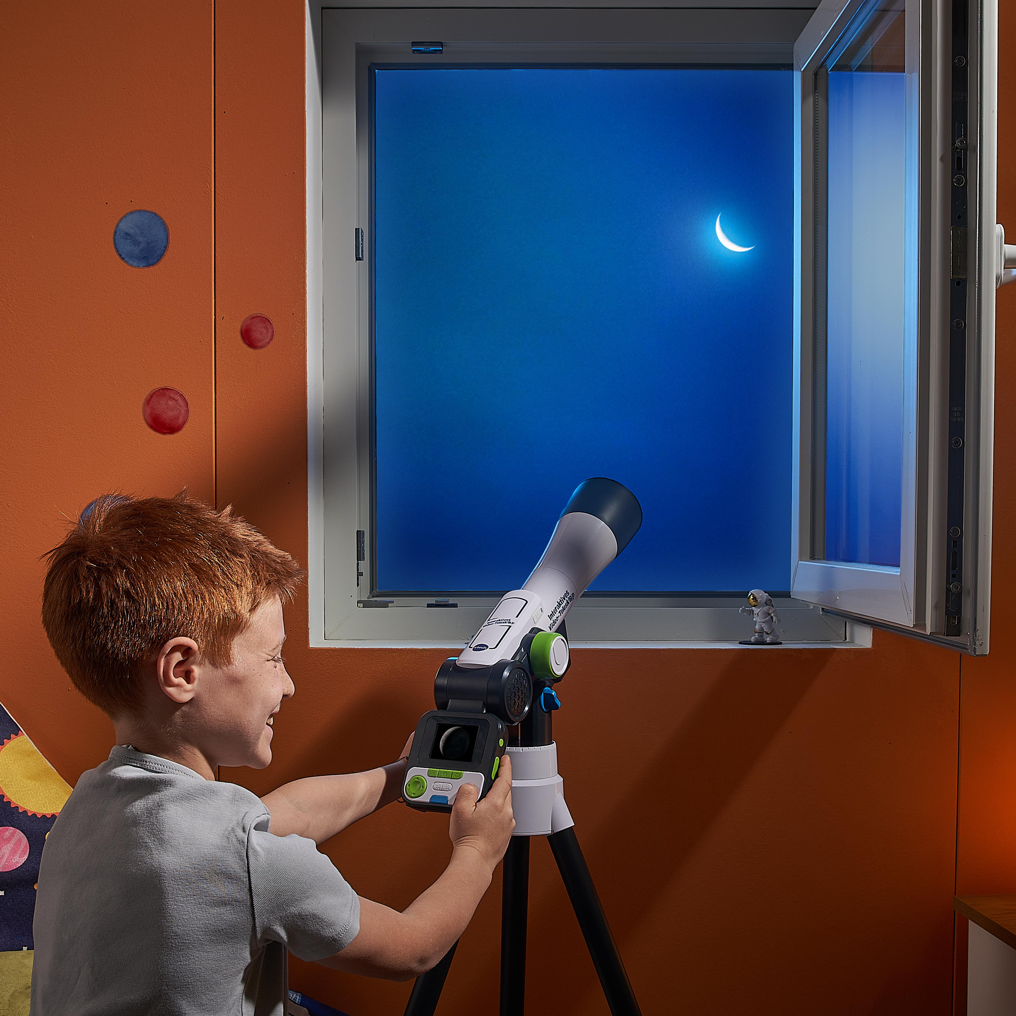 Junge schaut durch Teleskop auf Mondsichel im Fenster. Orange Wand, blauer Himmel. Spielzeug und Wanddeko.