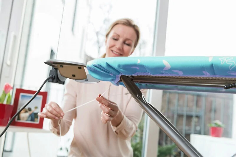 Kobieta dopasowuje pokrowiec deski do prasowania. W tle okno i ramka na zdjęcia.