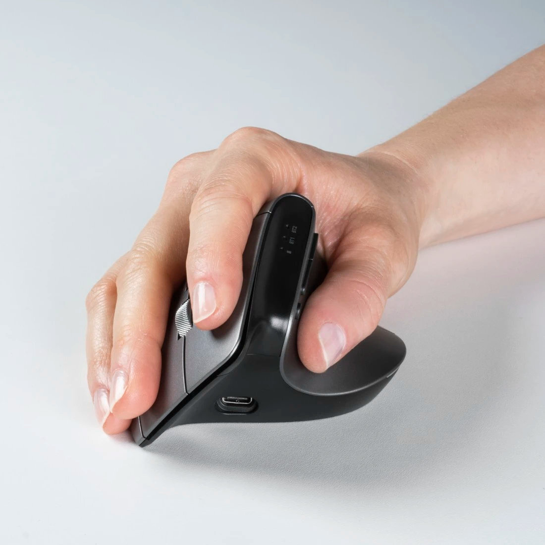 Dłoń trzymająca pionową ergonomiczną mysz. Jest ciemnoszara ze srebrnym kółkiem przewijania i portem ładowania.