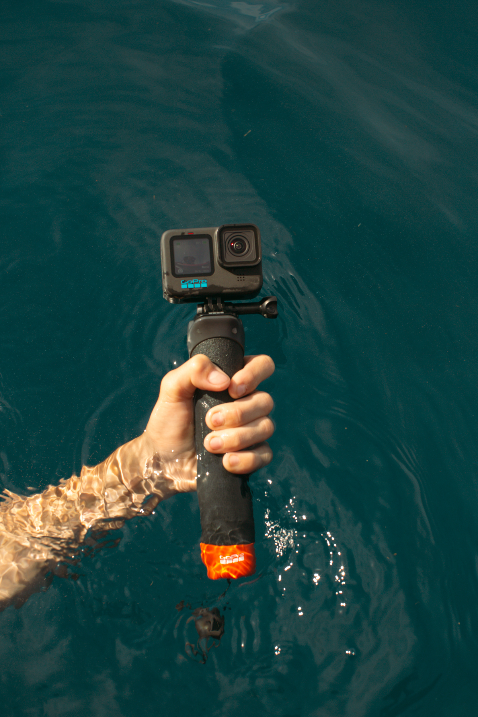 Eine Hand hält eine GoPro-Kamera an einem schwarzen Stab im blauen Wasser. Die Kamera zeigt zum Betrachter.