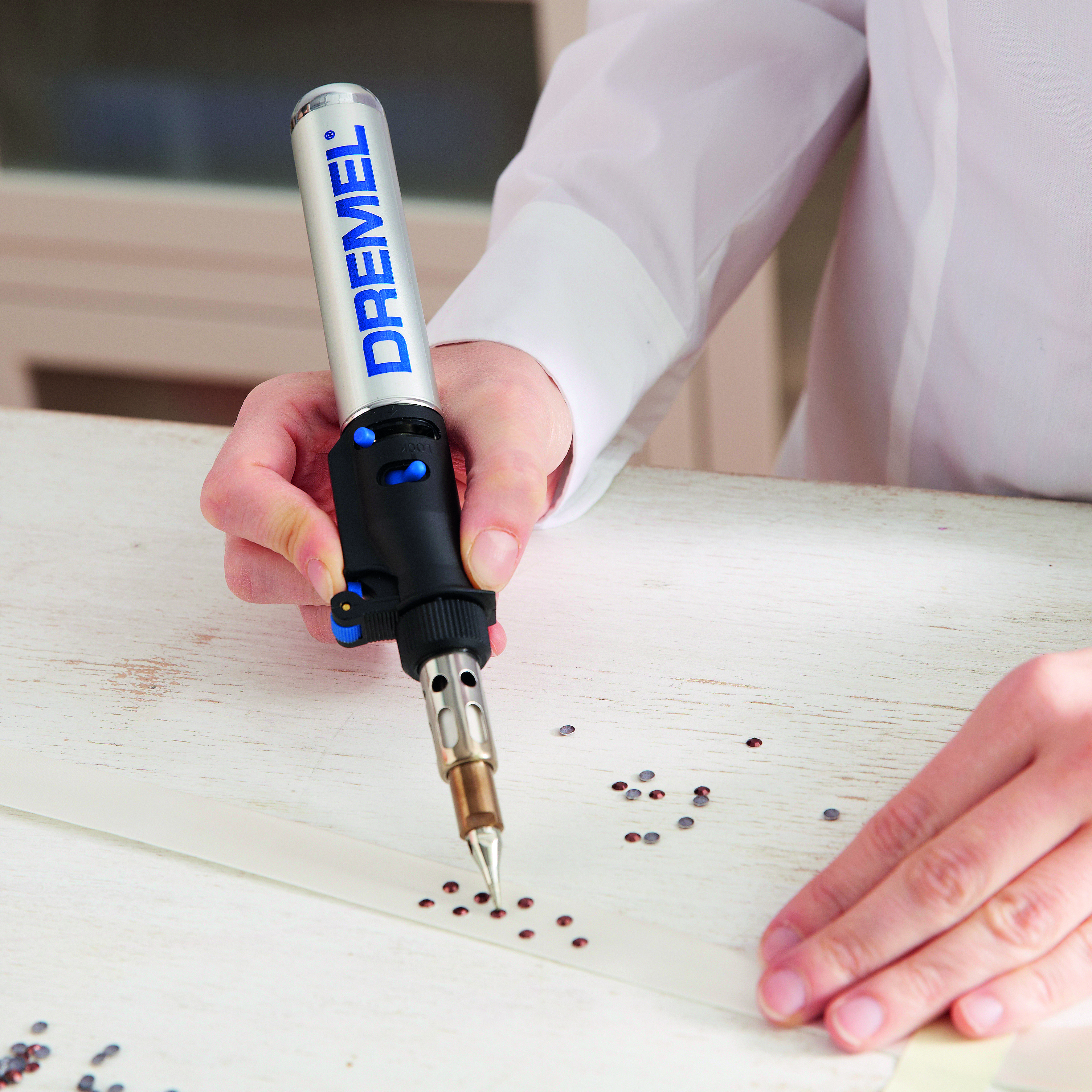 Egy személy Dremel szerszámot használ strasszkövek felvitelére fehér felületre, közeli felvétel.