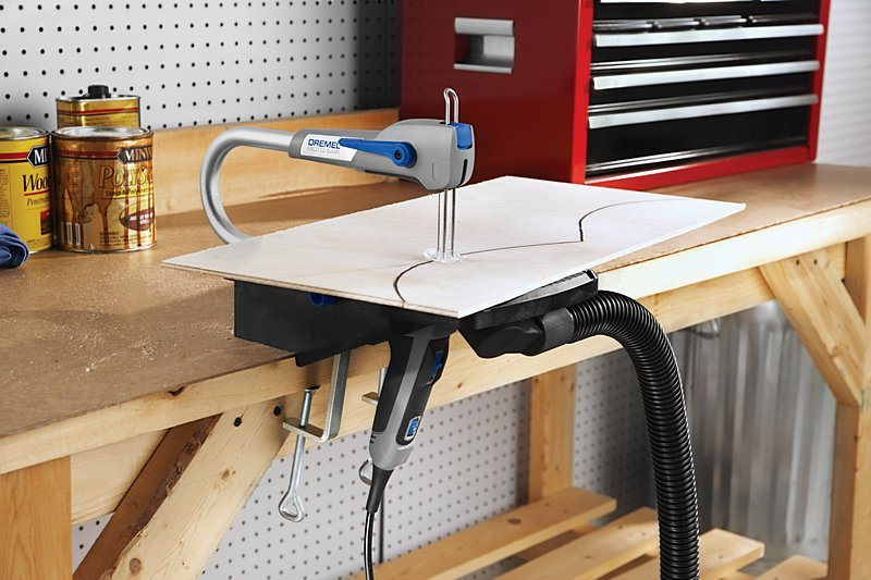 Dremel fűrész fát vág munkapadon, porszívó tömlővel.