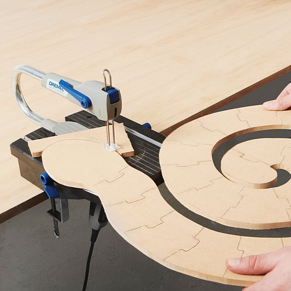 Egy Dremel szerszám fa puzzle darabot vág, két kézzel tartva egy asztalon.