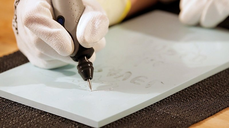 Egy személy forgó szerszámmal gravíroz egy világoskék felületet.