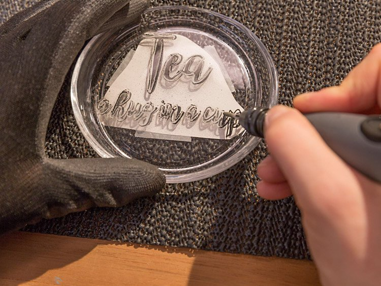 Személy szöveget gravíroz egy átlátszó poháralátétre. A szöveg a következő: 'Tea, ölelés egy csészében'.