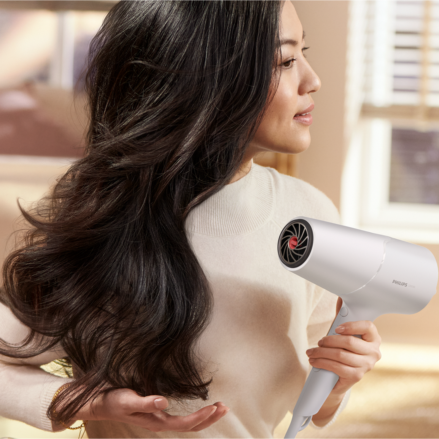 A woman holds a white hairdryer to her hair. The background is slightly blurred.