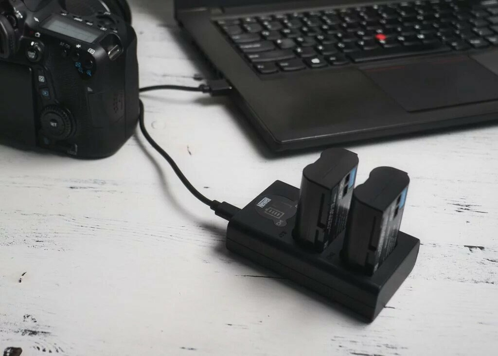 Aparat i laptop z podłączoną ładowarką. Baterie aparatu znajdują się w ładowarce. Ekran laptopa nie jest widoczny.