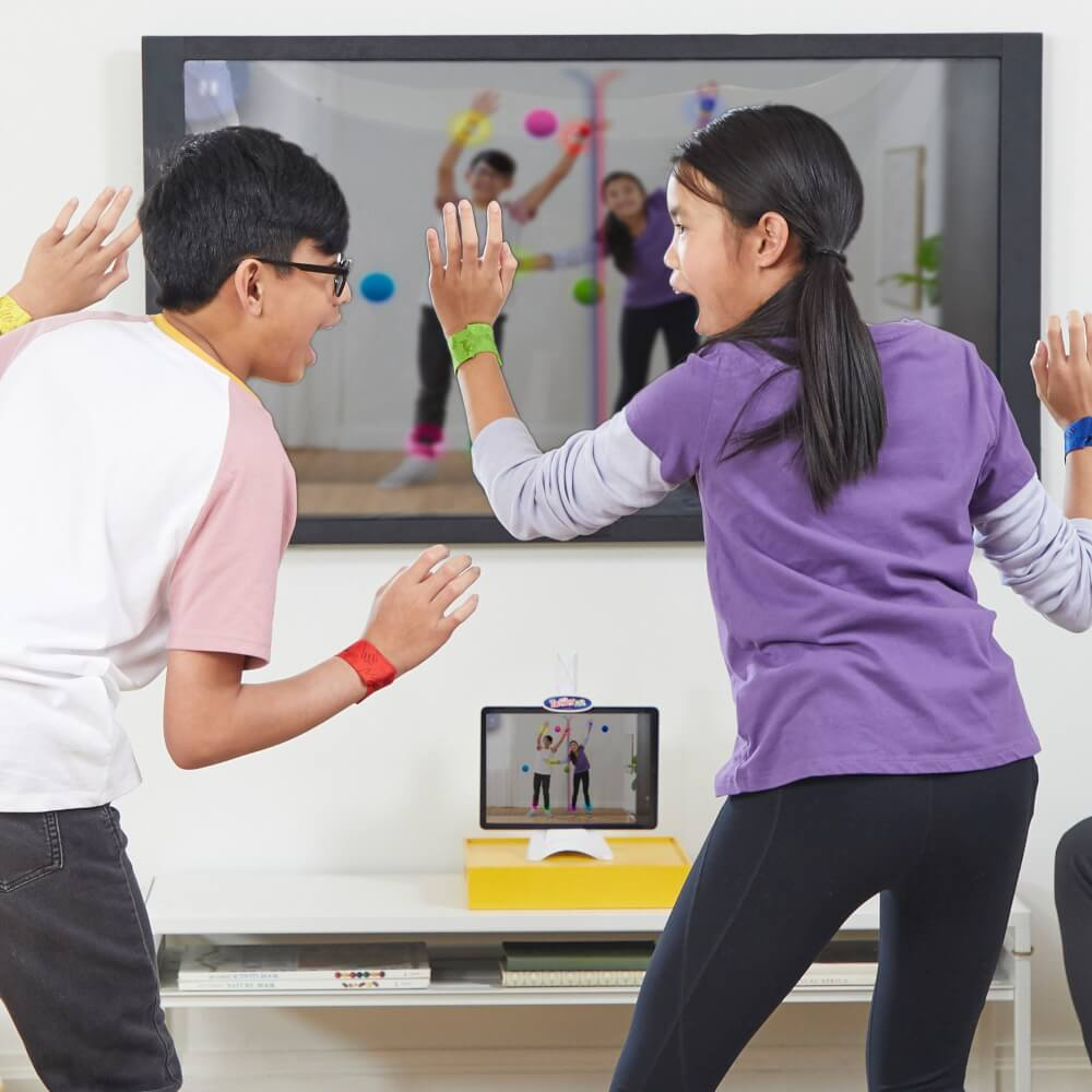 Zwei Kinder spielen ein Spiel mit einem Tablet, mit einem großen Bildschirm, der sie und bunte Bälle in einem hellen Raum zeigt.