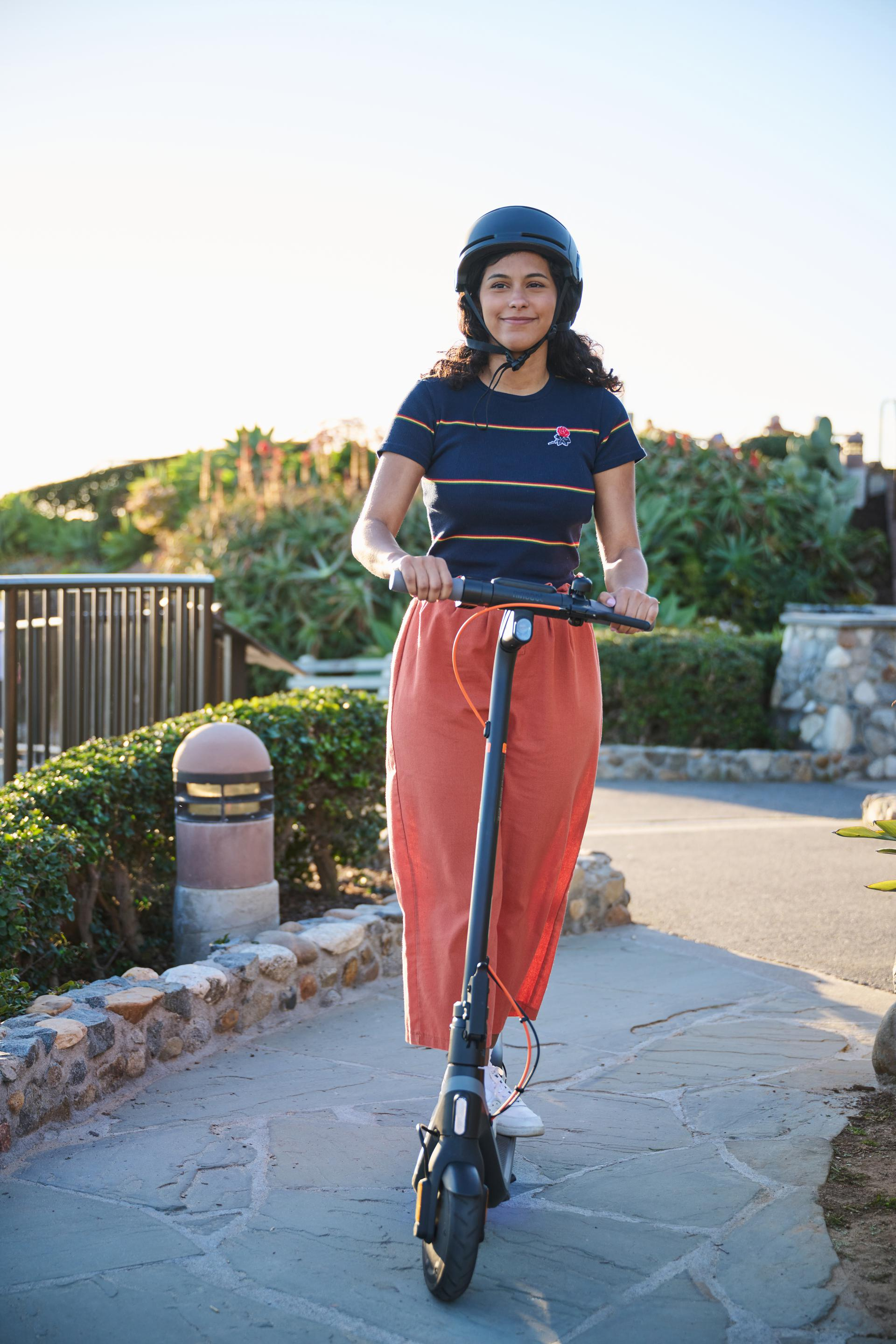 Una mujer en un patinete eléctrico, con casco, en un camino pavimentado.