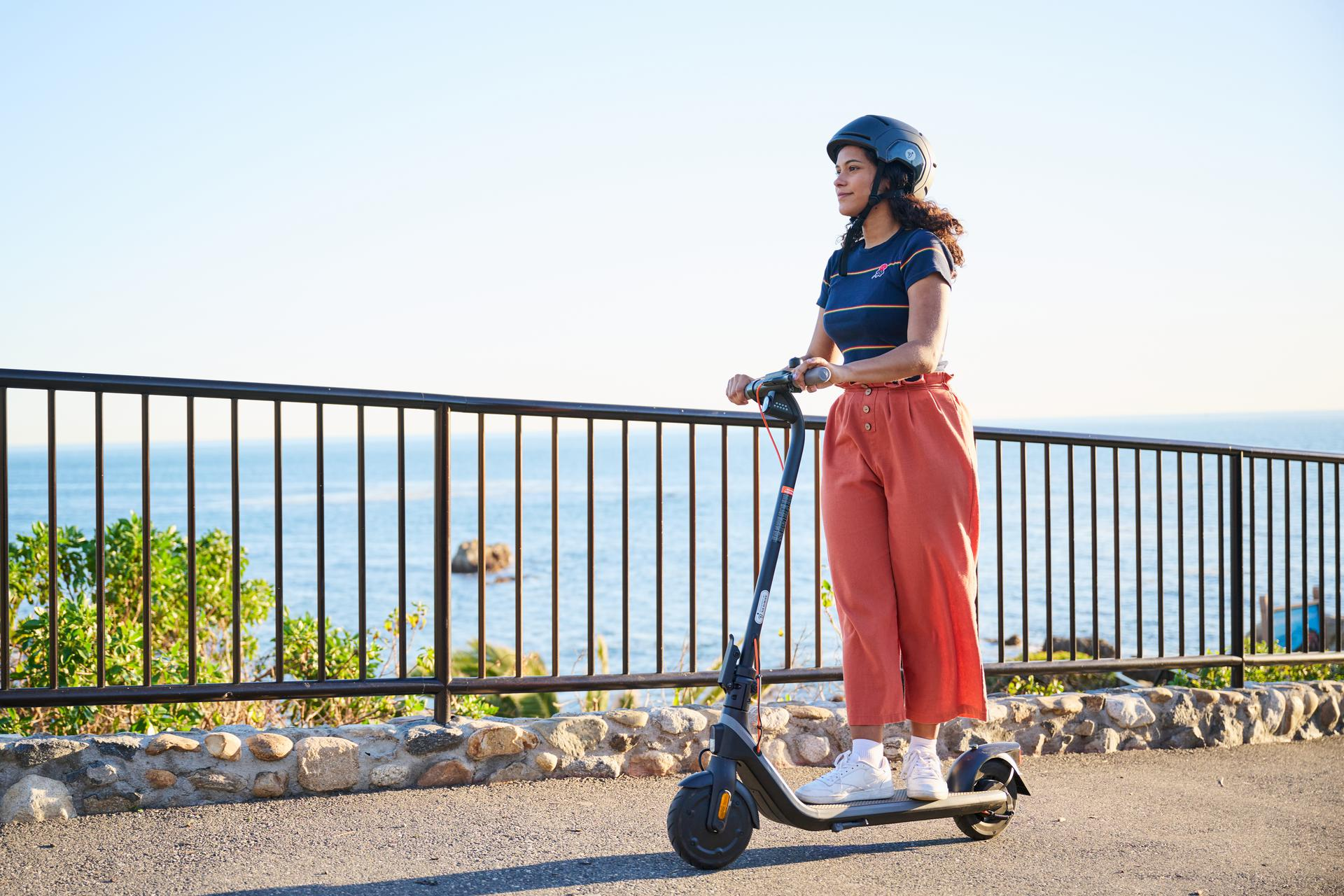 Una mujer en un patinete eléctrico, con casco, montando por un camino costero.