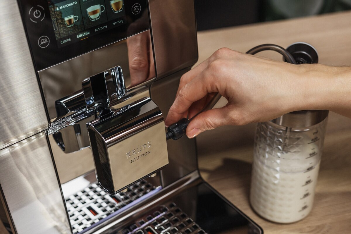 Osoba reguluje pokrętło na srebrnym ekspresie do kawy. Pojemnik na mleko po prawej.