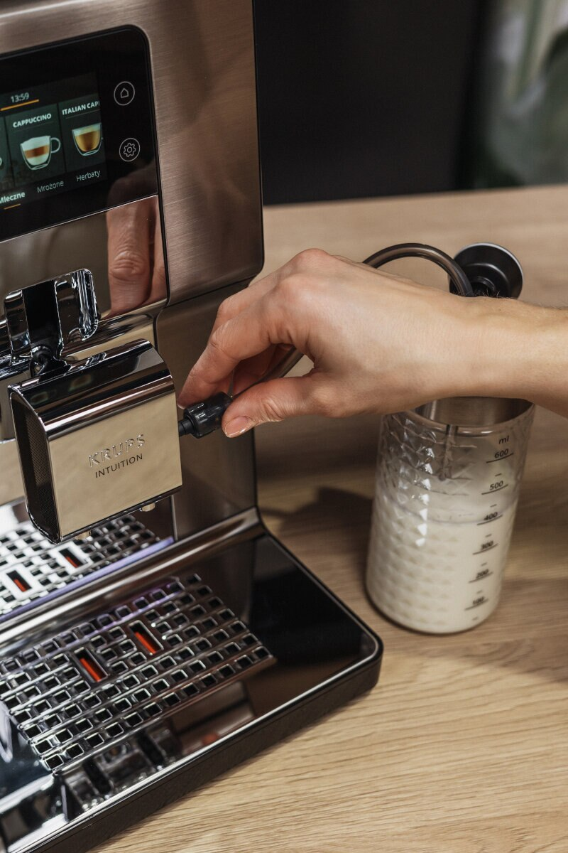 Ręka używająca ekspresu do kawy Krups Intuition, nalewająca mleko do szklanki.