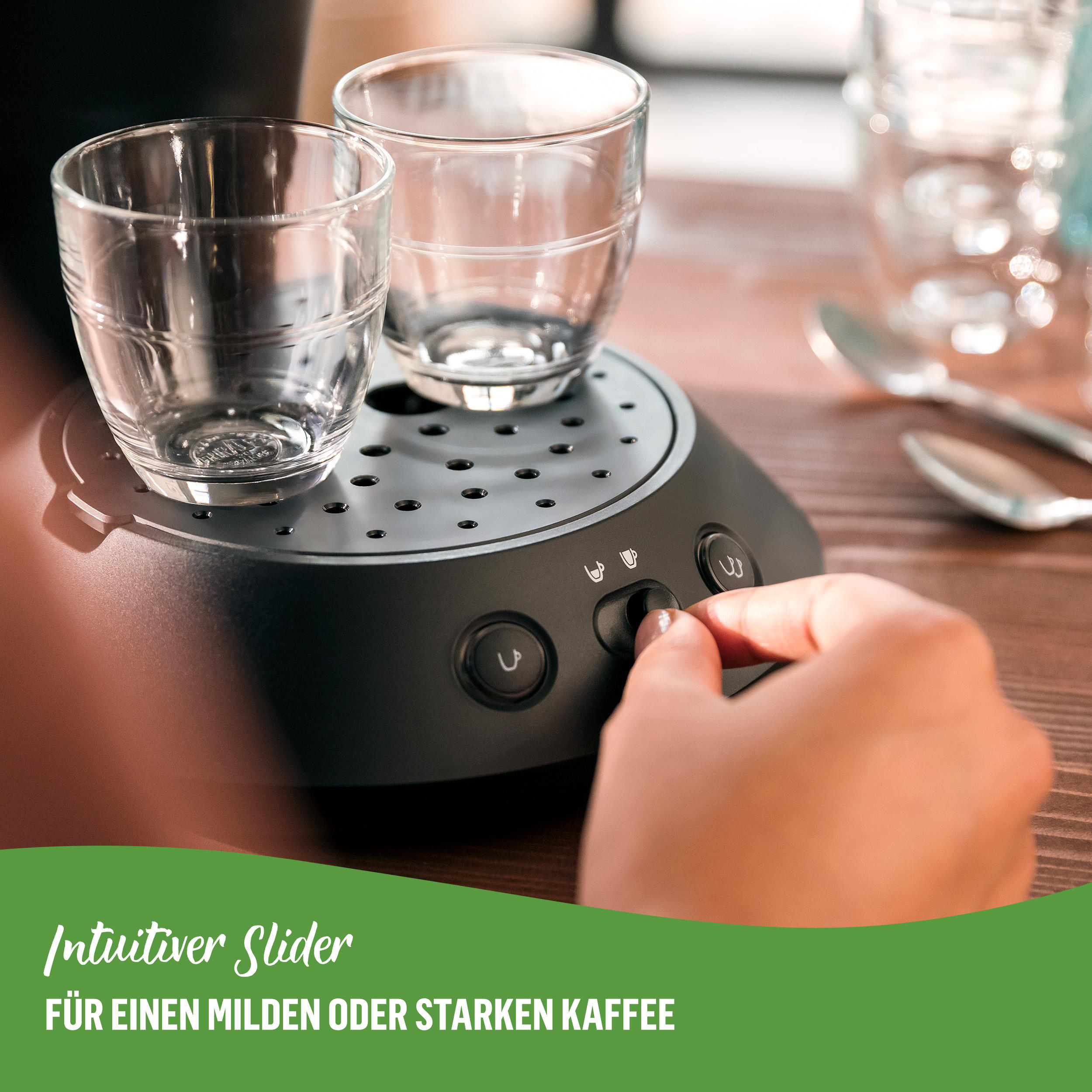 Zwei leere Gläser auf einer schwarzen Kaffeemaschine. Eine Hand bedient einen Steuerdrehknopf. Holztisch.