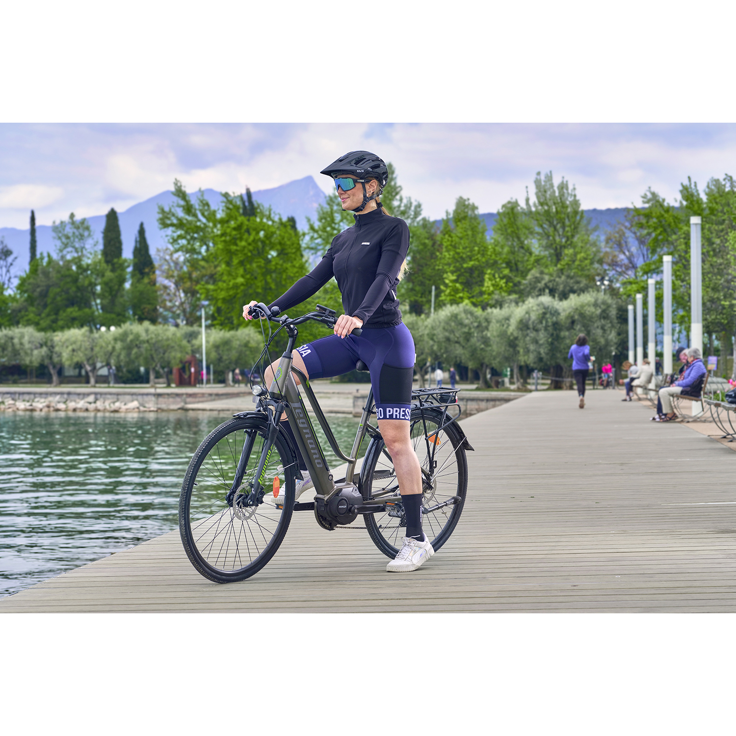 Donna su una bici nera in riva al lago. Indossa casco, occhiali da sole e abbigliamento sportivo.