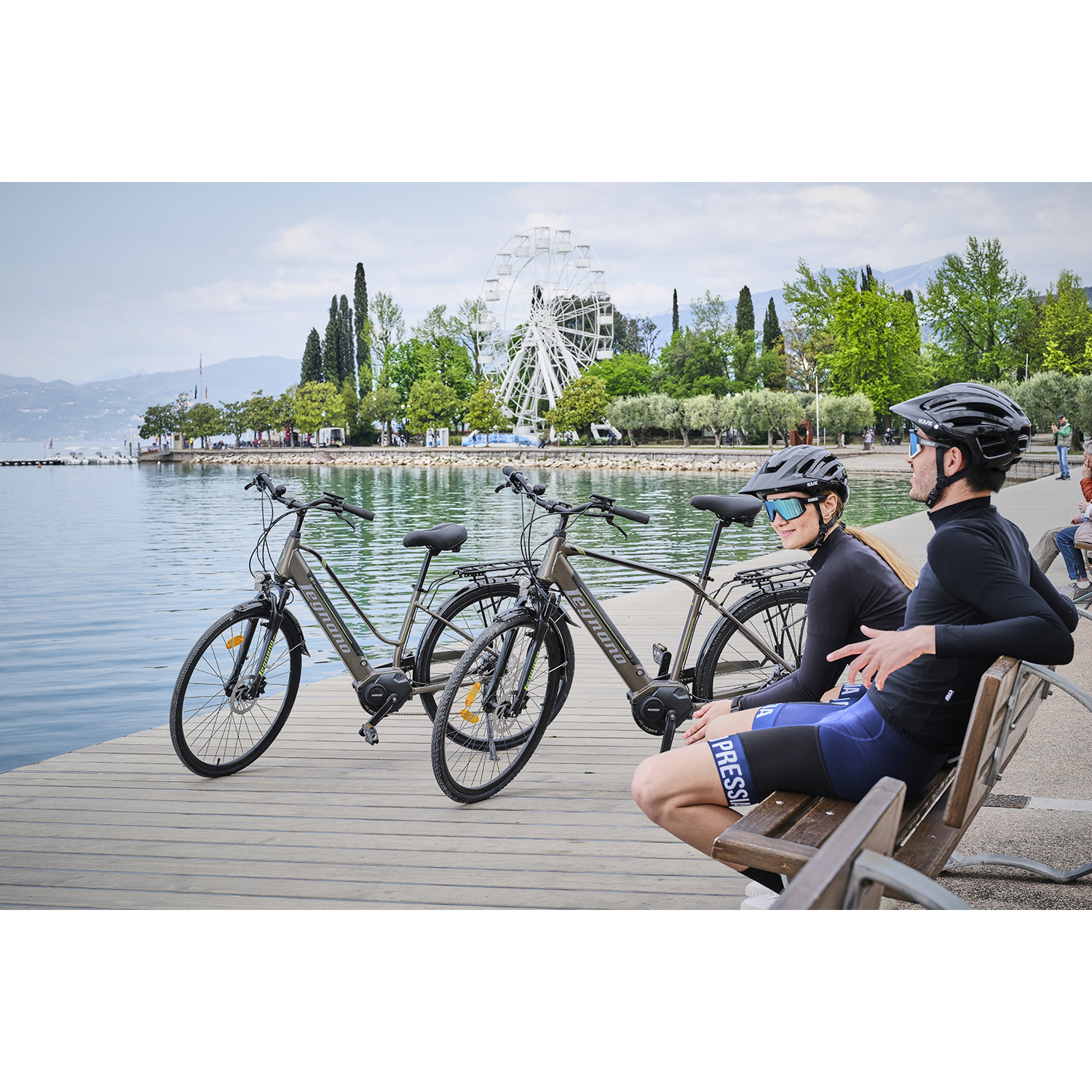 Due bici elettriche su un molo, una coppia seduta vicino, lago e montagne sullo sfondo.