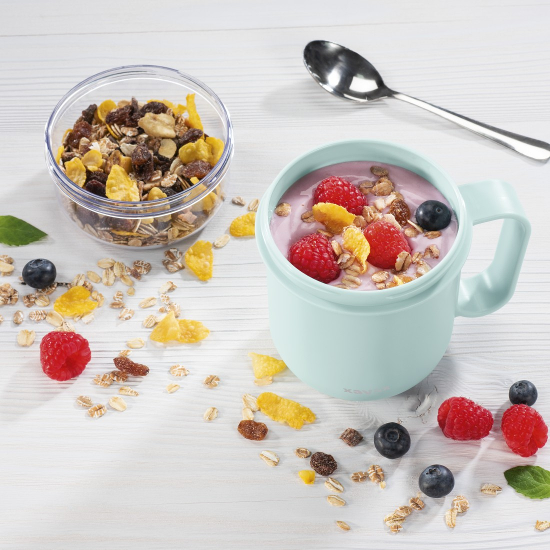 Un bol de granola et une tasse avec du yaourt, des framboises et des myrtilles. Une cuillère à côté.