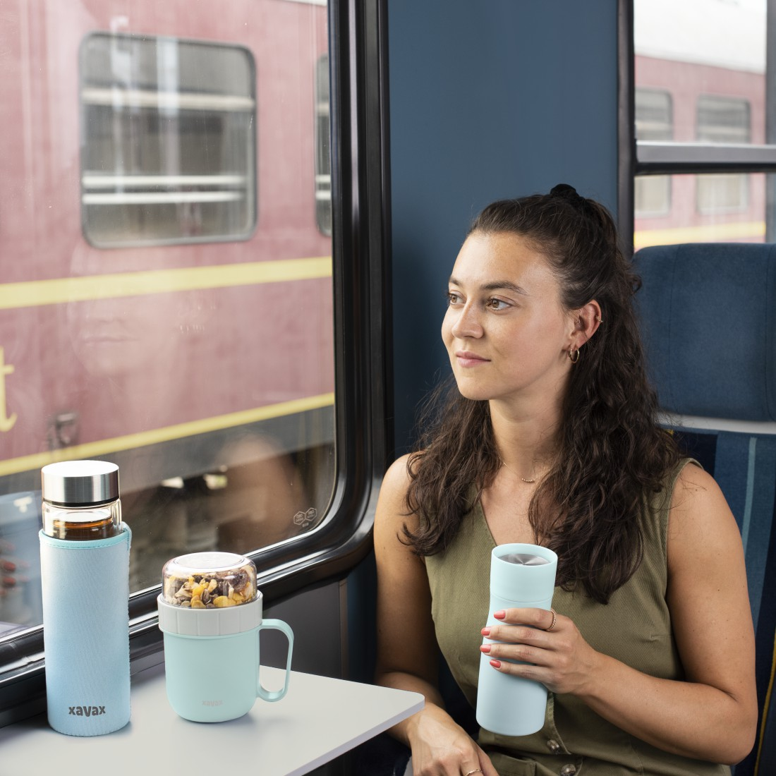 Femme dans un train regardant par la fenêtre, avec des bouteilles d'eau et un récipient à collation sur la table.