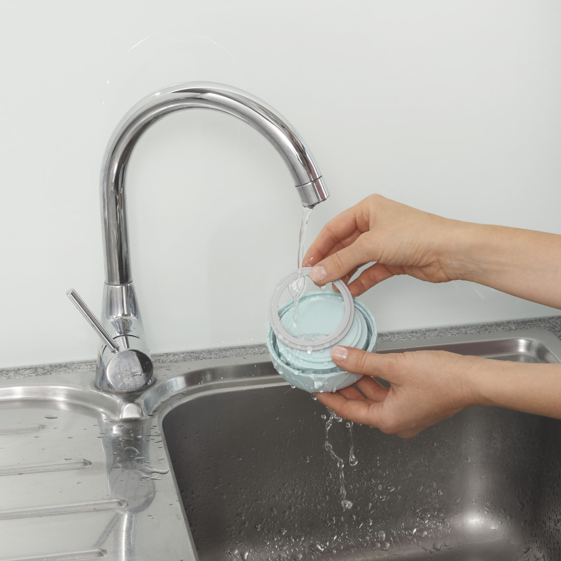 Mains lavant un récipient bleu et gris sous un robinet de cuisine en acier inoxydable.