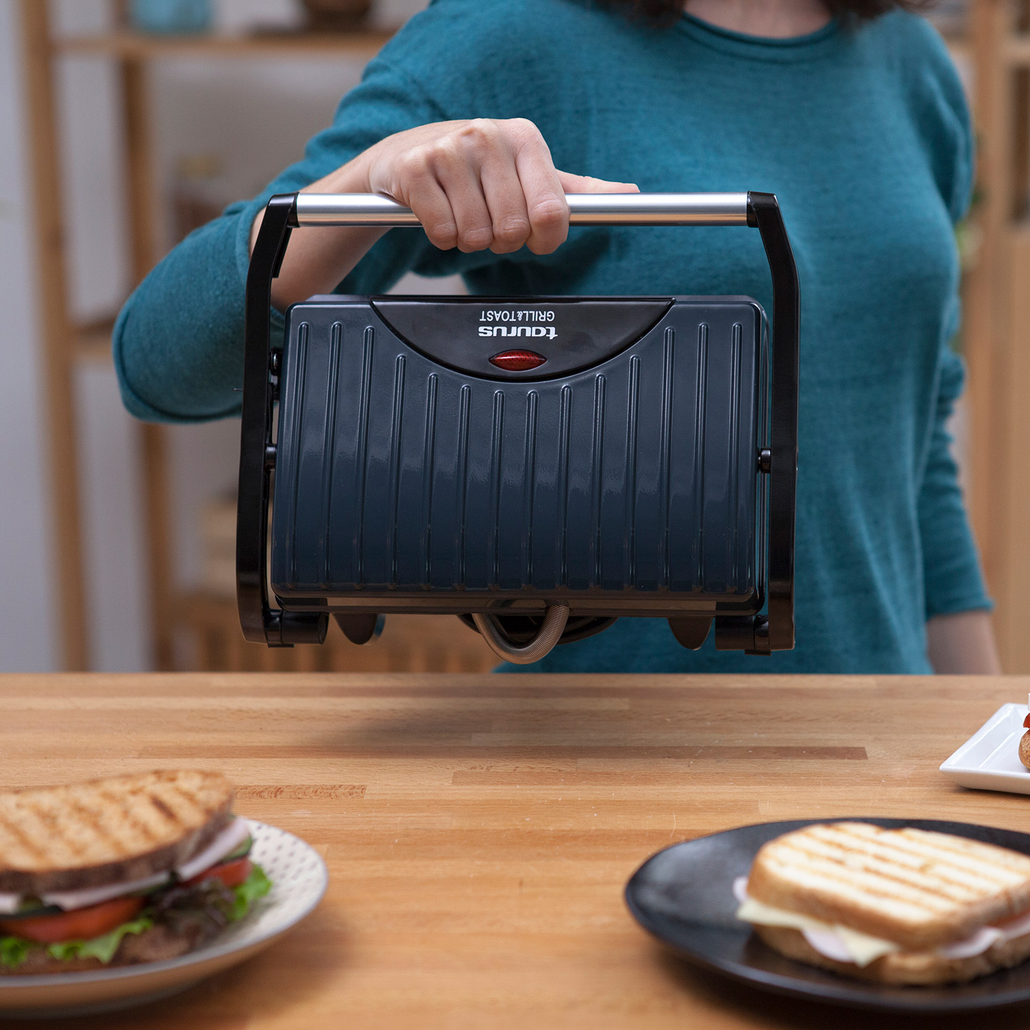 Persona sosteniendo una parrilla negra. Dos sándwiches en primer plano en mesa madera.