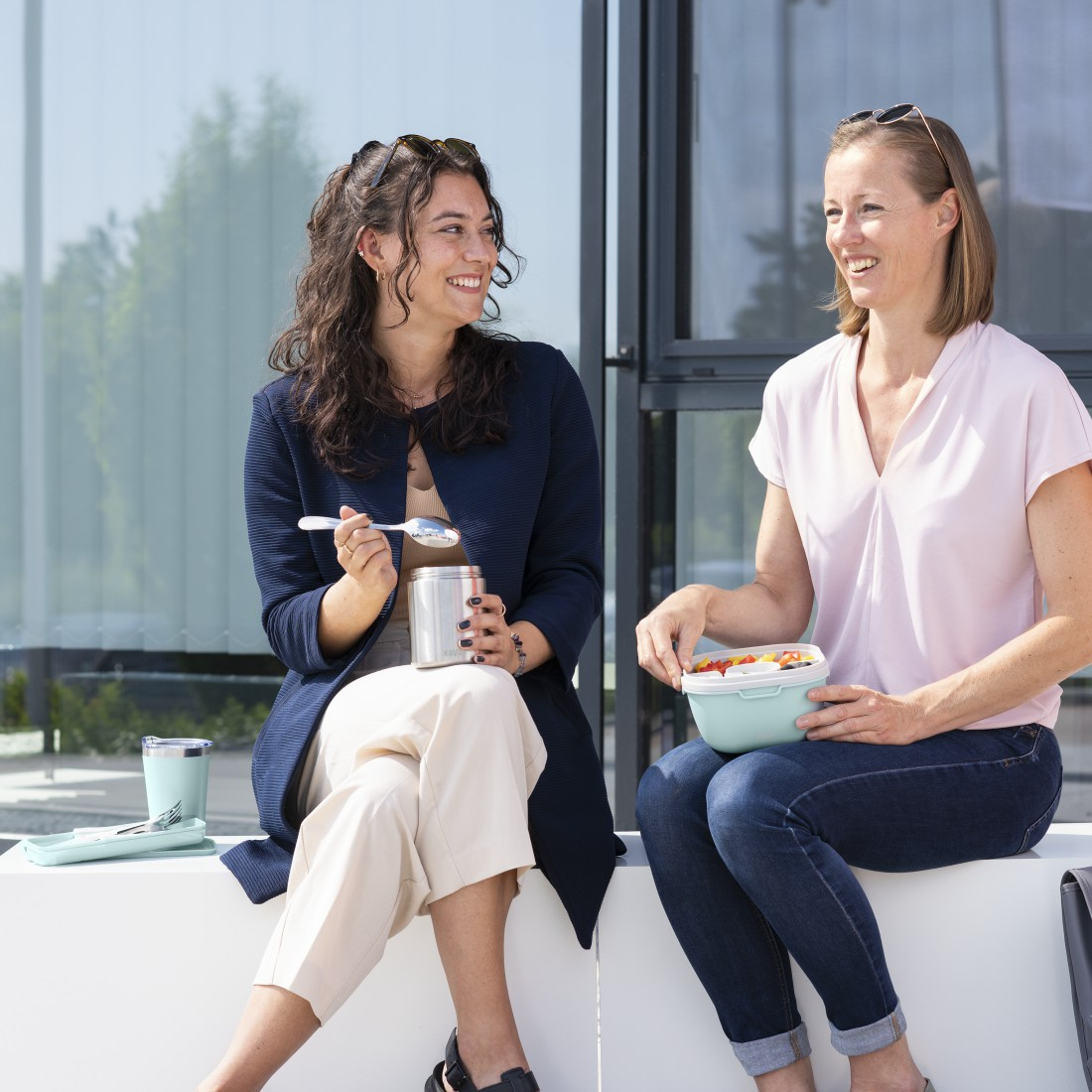Deux femmes souriantes, assises dehors, l'une tenant un récipient, l'autre une boîte à lunch.