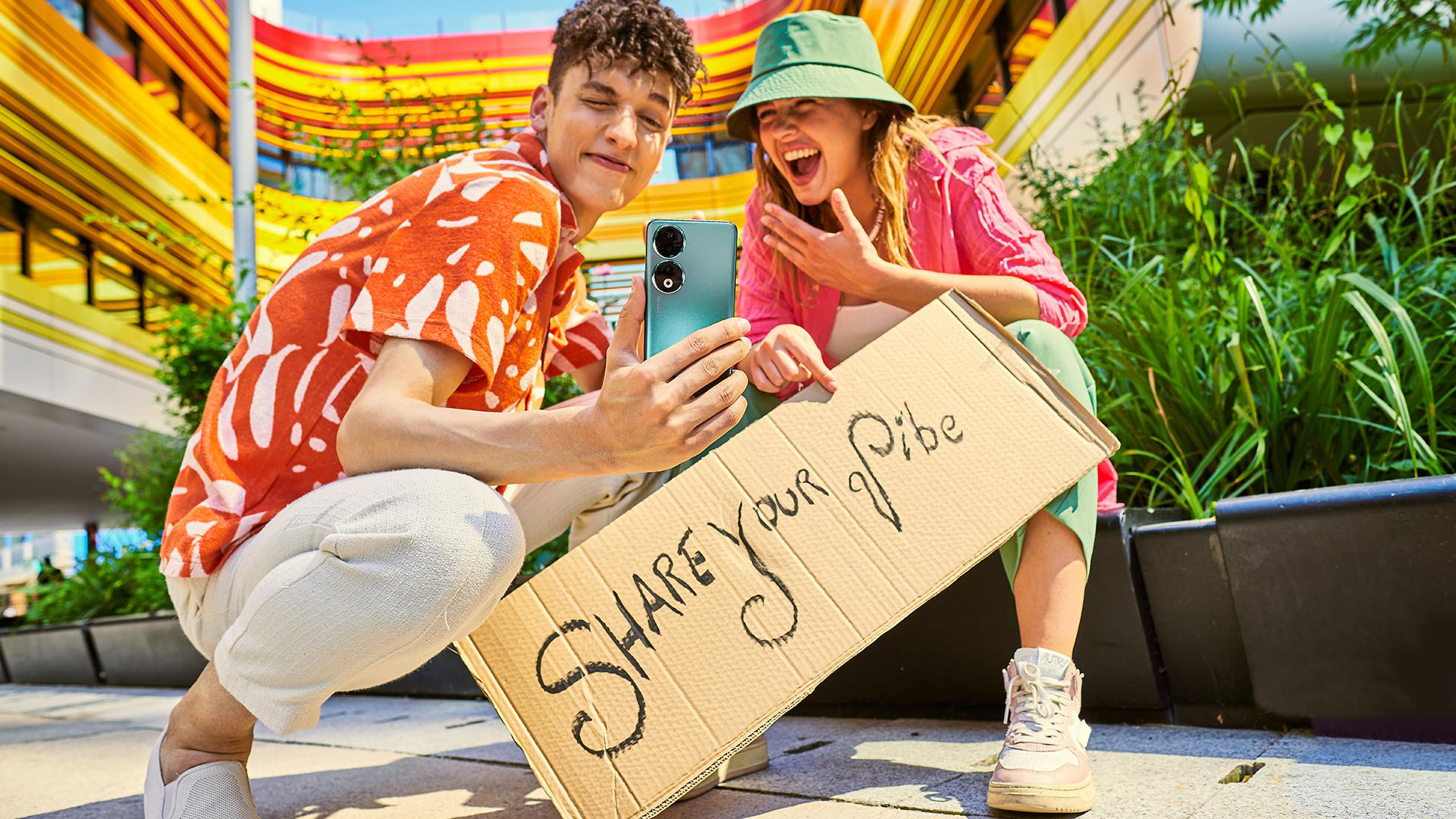 Zwei Personen machen ein Selfie, halten ein Telefon, mit einem Schild mit der Aufschrift 'Share your vibe'.