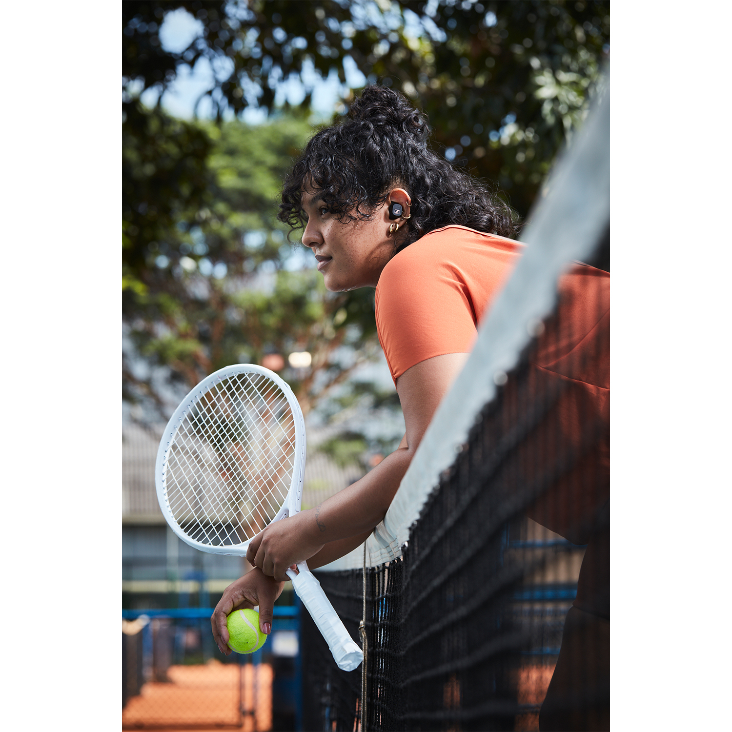 Donna in maglietta arancione con cuffie, tiene una racchetta e una palla da tennis su un campo.