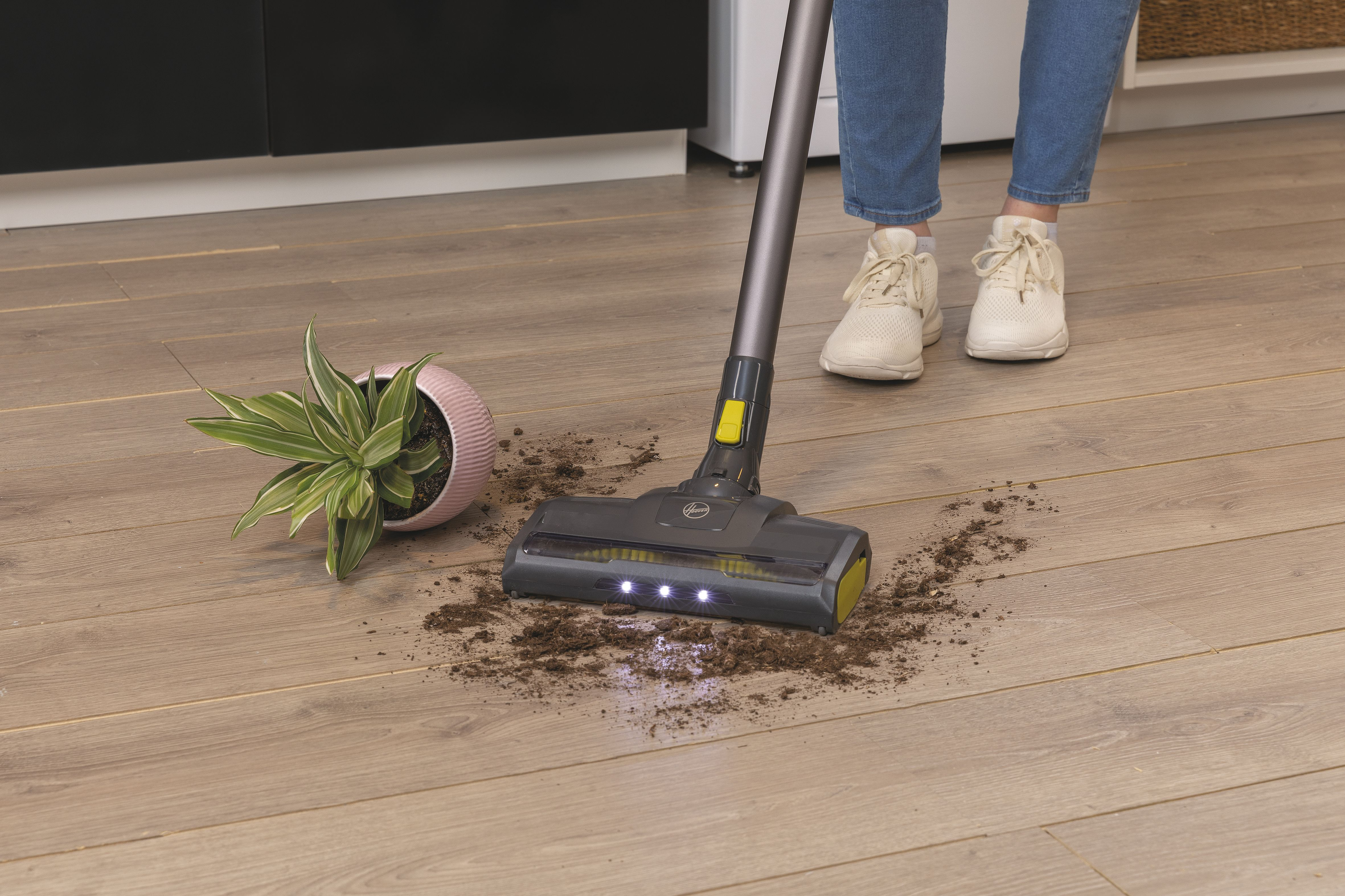 Una aspiradora en suelo de madera, limpiando suciedad. Una planta a la izquierda, pies visibles.