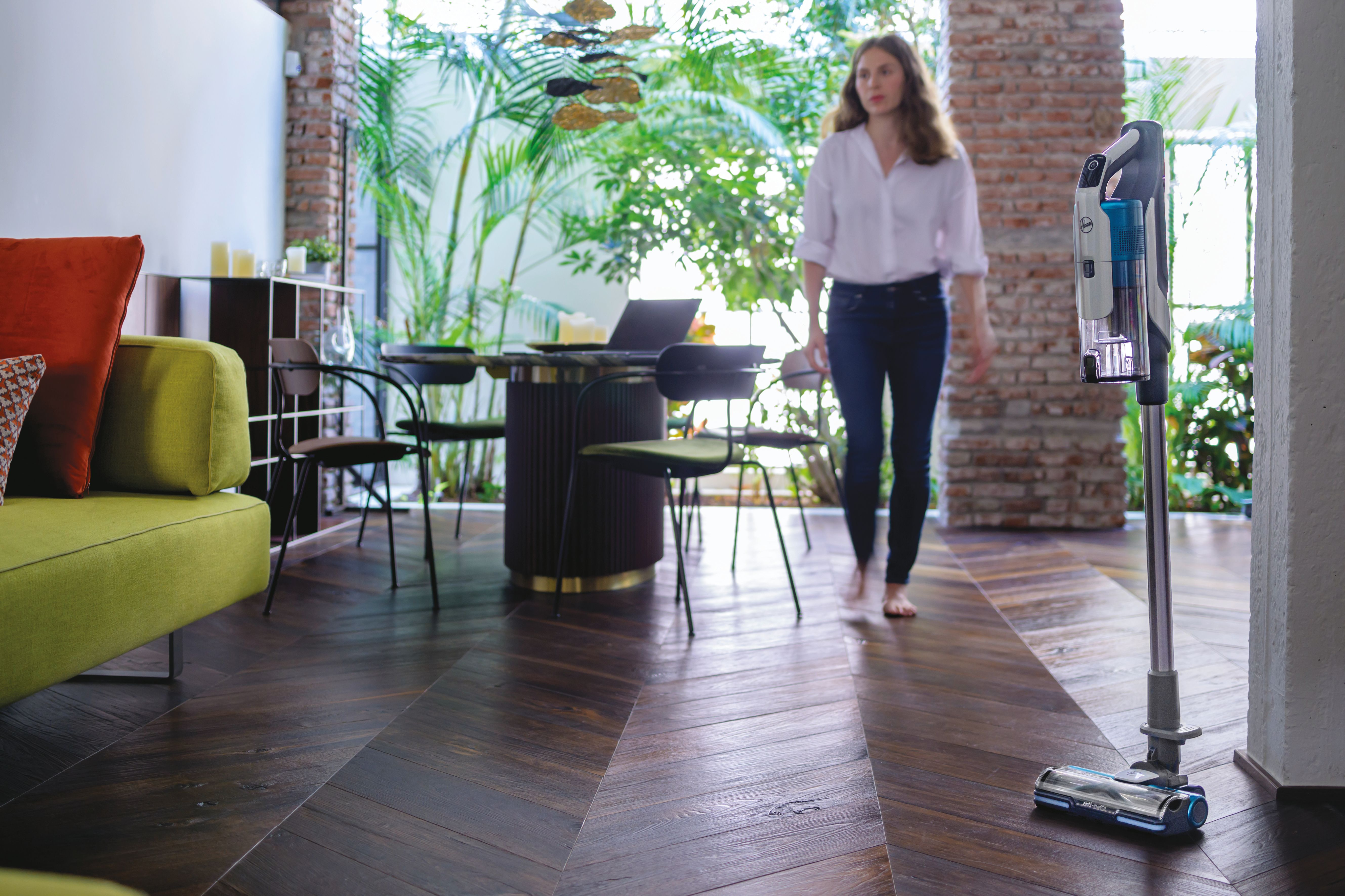 Una mujer caminando en una sala de estar moderna, una aspiradora está en el fondo.