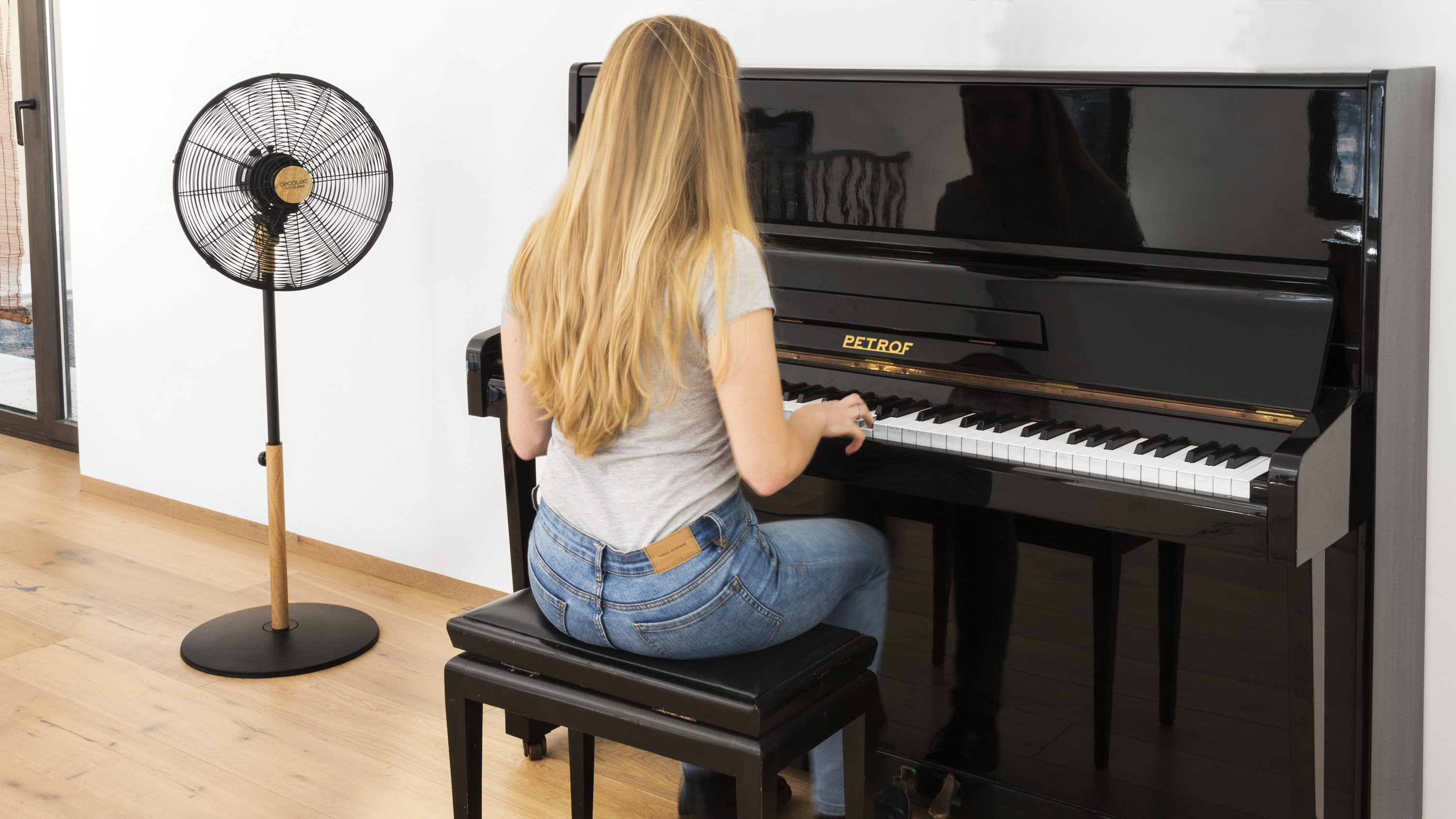 Una mujer toca el piano. Un piano negro y un ventilador están en la habitación. El ventilador está a la izquierda.