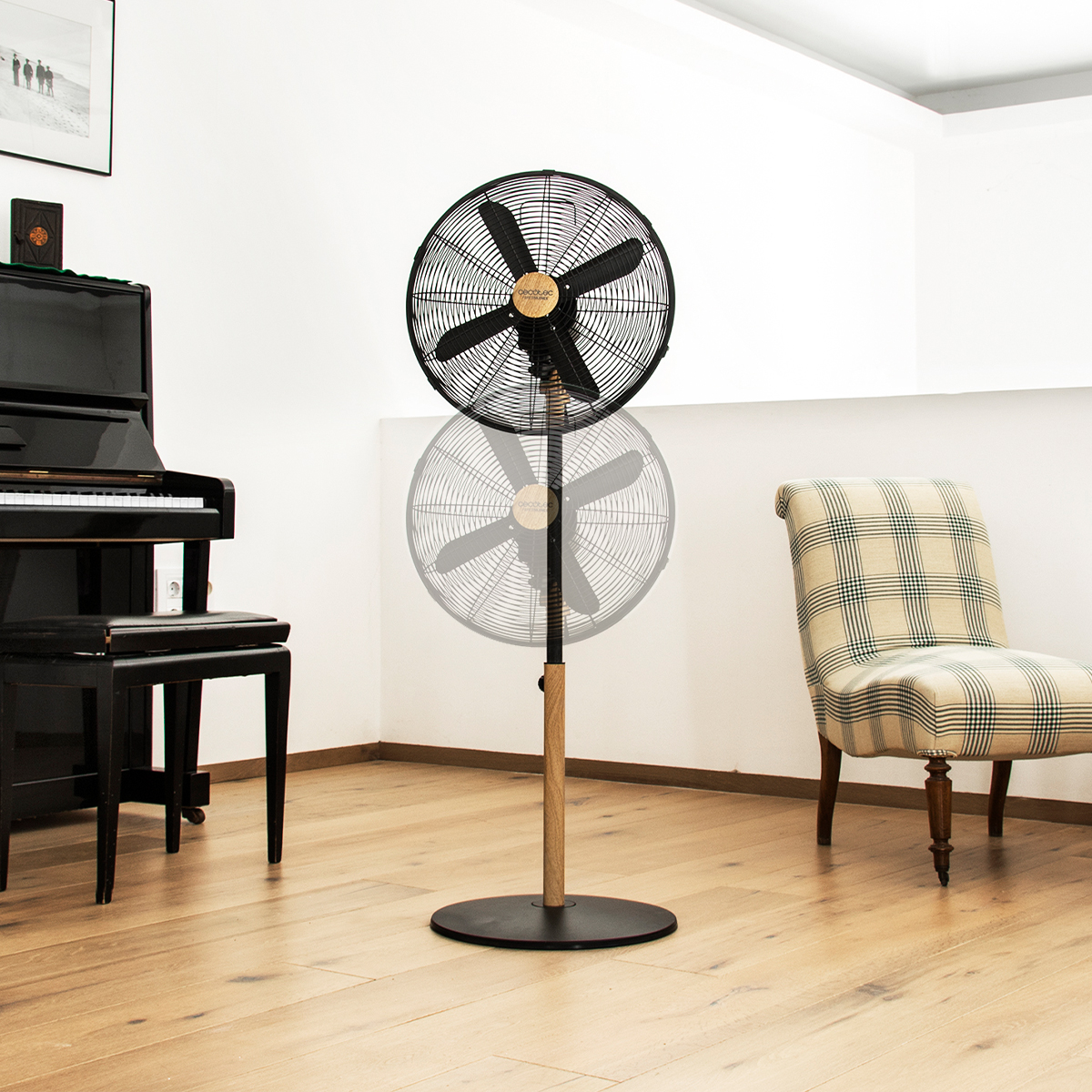 Un ventilador de pie en una habitación. Aspas negras, soporte de madera y negro. Piano y silla en el fondo.