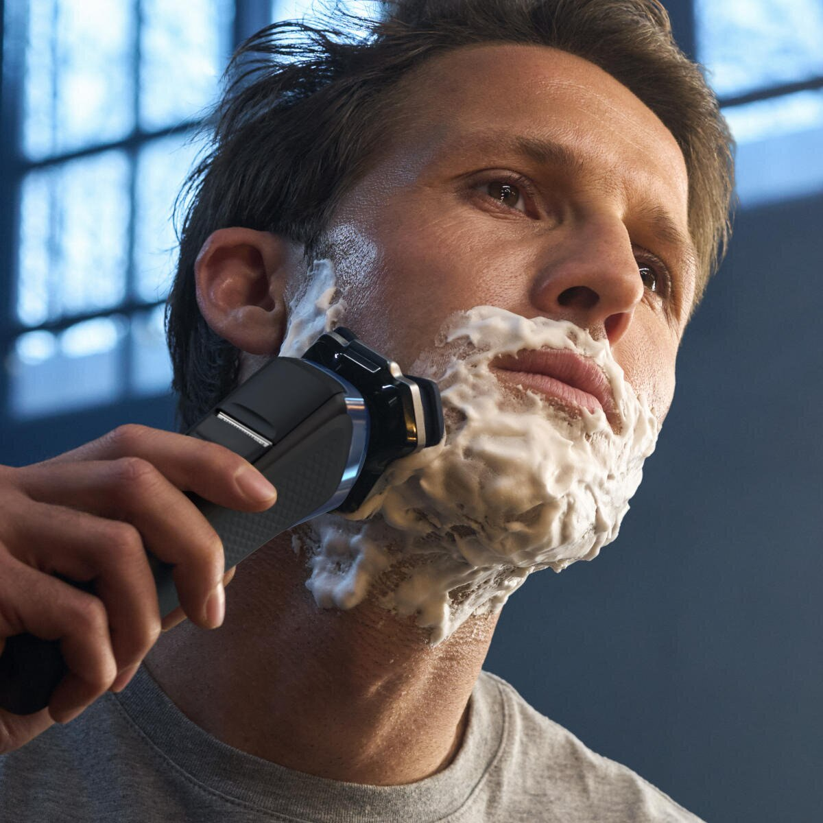 Mężczyzna goli się maszynką elektryczną, biała pianka na twarzy. Ciemne tło.