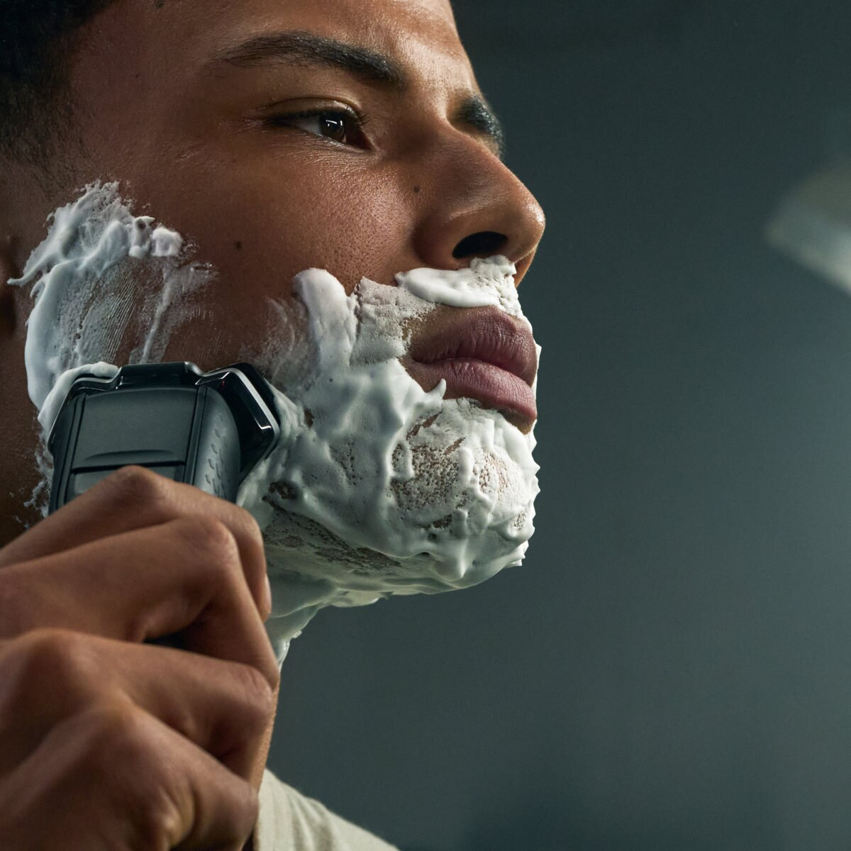 Mężczyzna goli twarz golarką elektryczną, nakładając krem do golenia. Zbliżenie, ciemne tło.