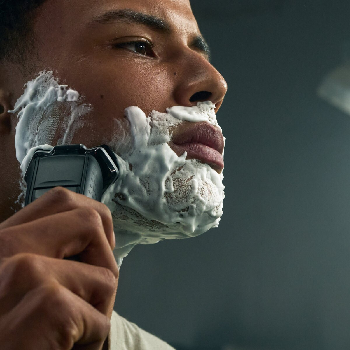 Mężczyzna goli twarz czarną golarką elektryczną. Biała pianka do golenia pokrywa jego twarz.