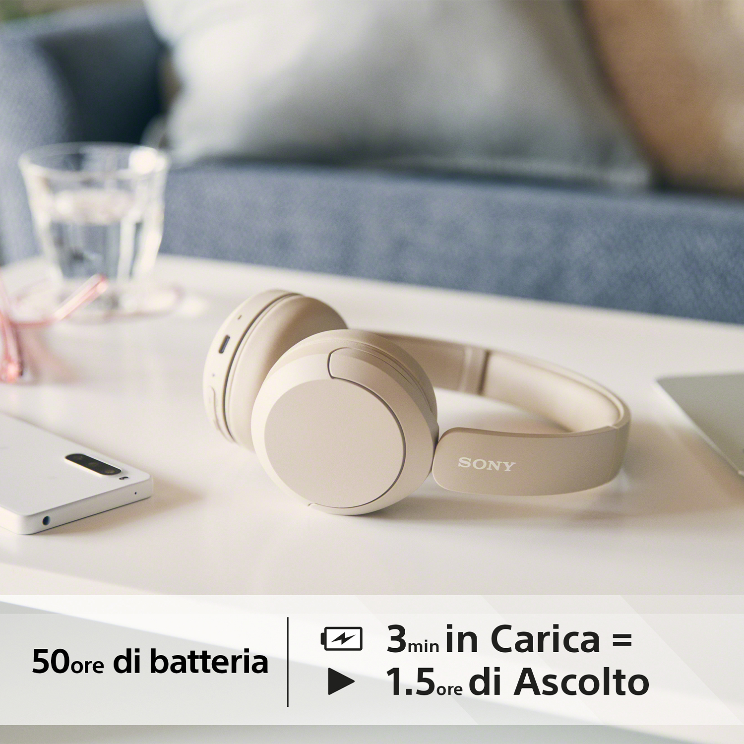 Cuffie Sony beige su un tavolo. Un telefono e un bicchiere sono anche sul tavolo. È presente del testo.