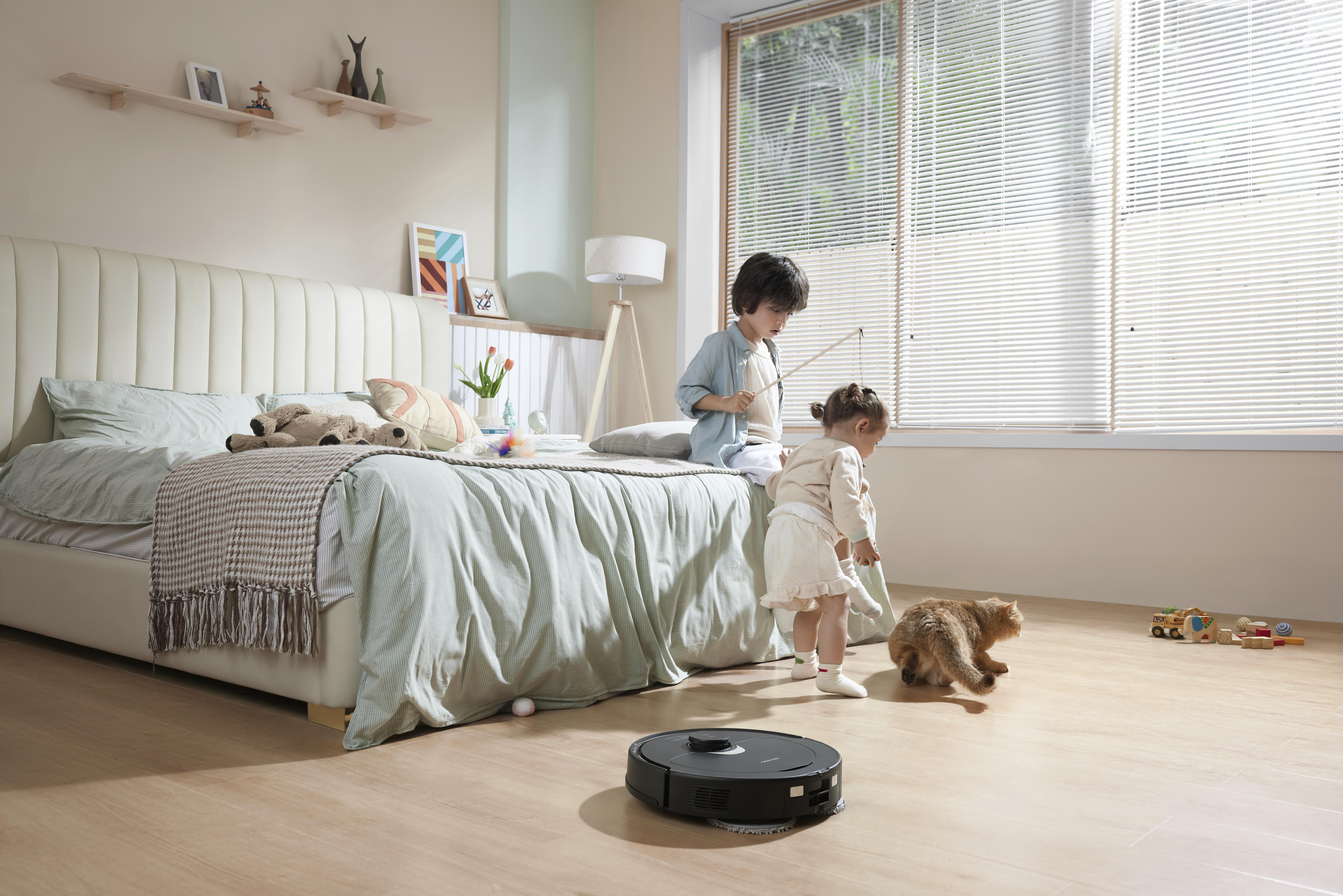 Zwei Kinder spielen auf einem Bett mit Katze und Roboter-Staubsauger auf dem Boden.