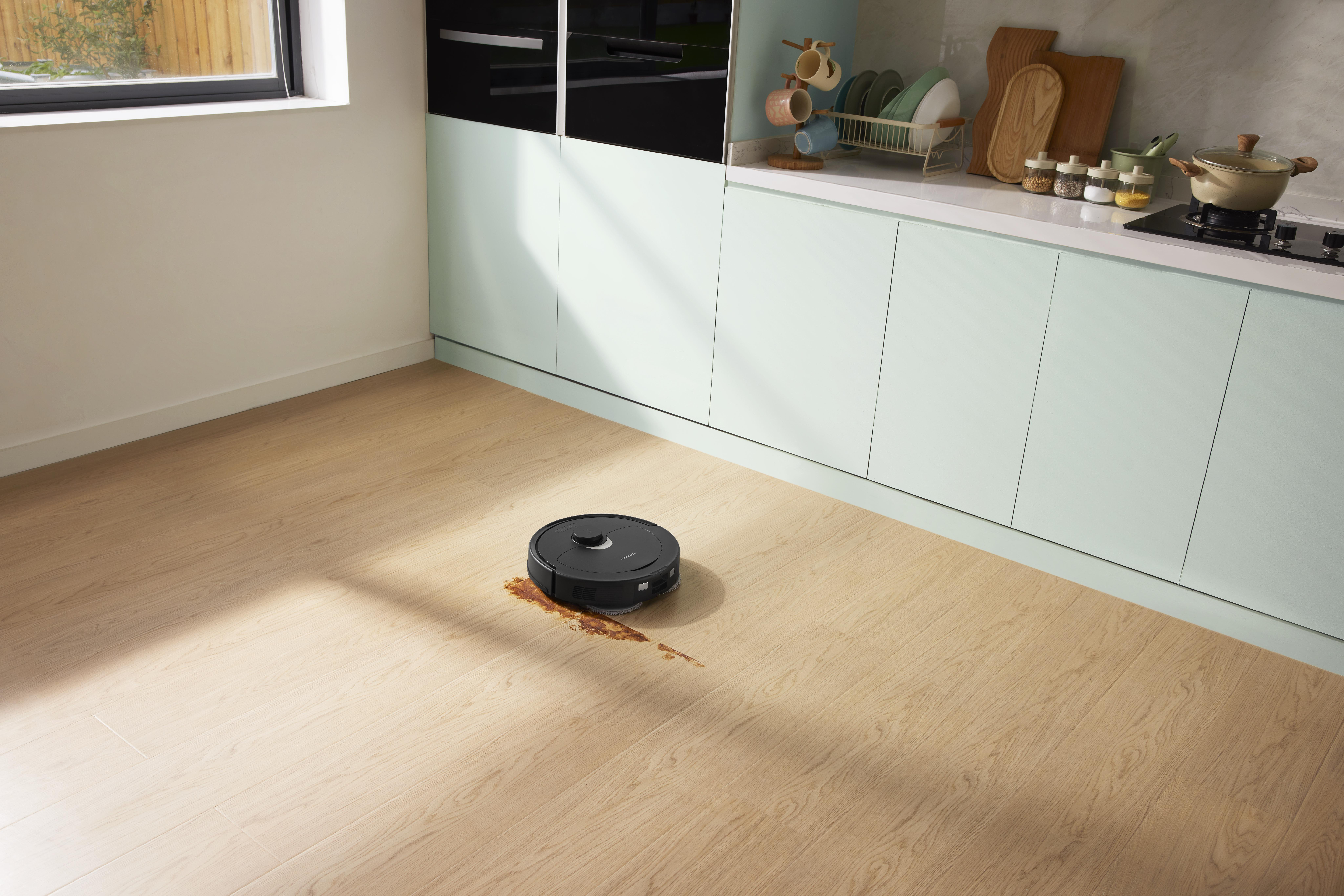 Ein Roboter-Staubsauger auf Holzboden mit Verschüttung. Küchenschränke und Geräte im Hintergrund.