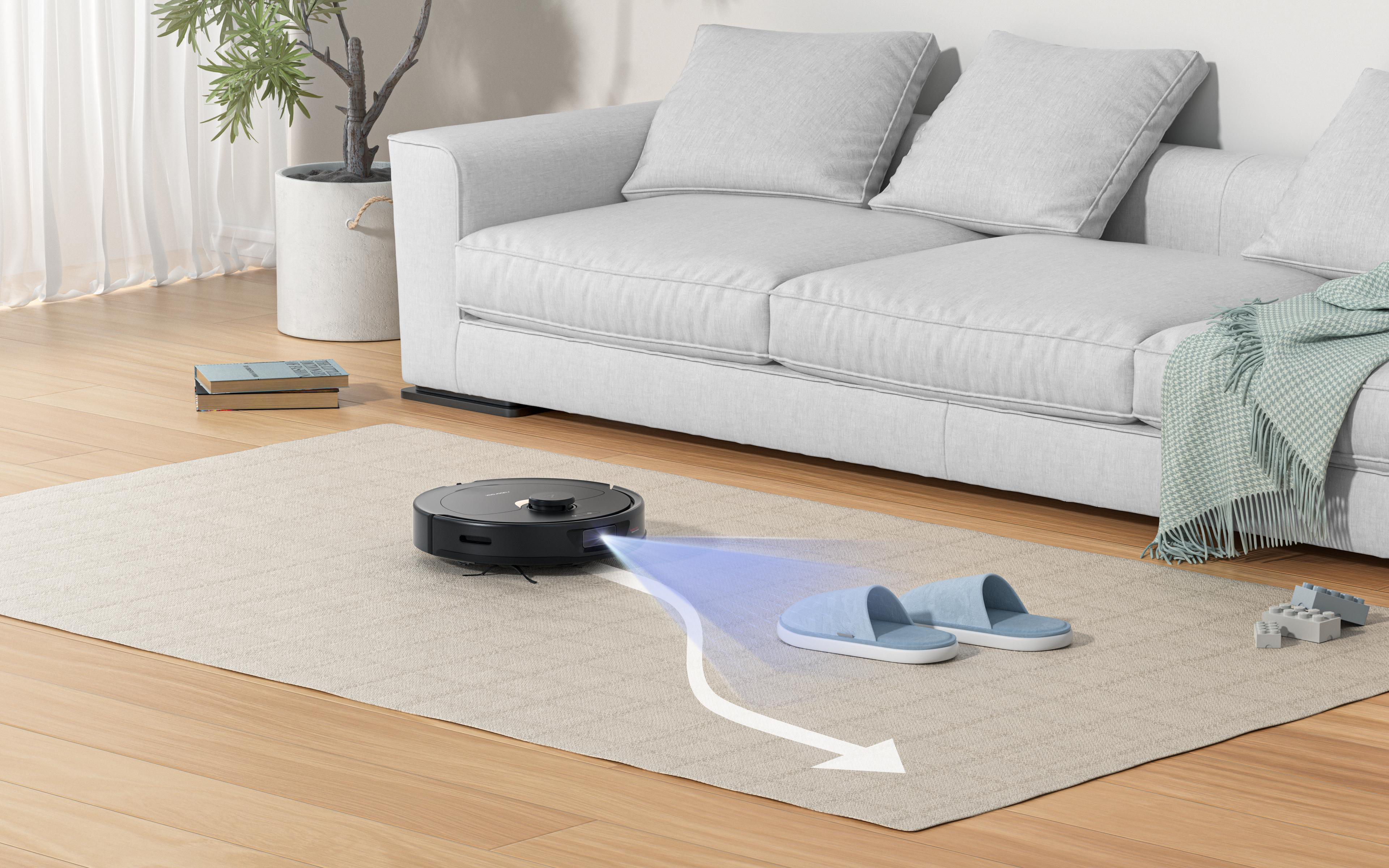 Ein Roboter-Staubsauger auf einem Teppich beim Staubsaugen. Ein Paar blaue Hausschuhe und ein kleines Spielzeug sind vorhanden.