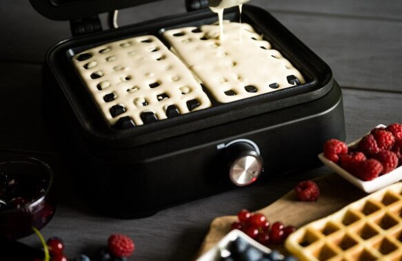 Gofry robione w gofrownicy. W pobliżu składniki takie jak jagody, dżem i inne dodatki.