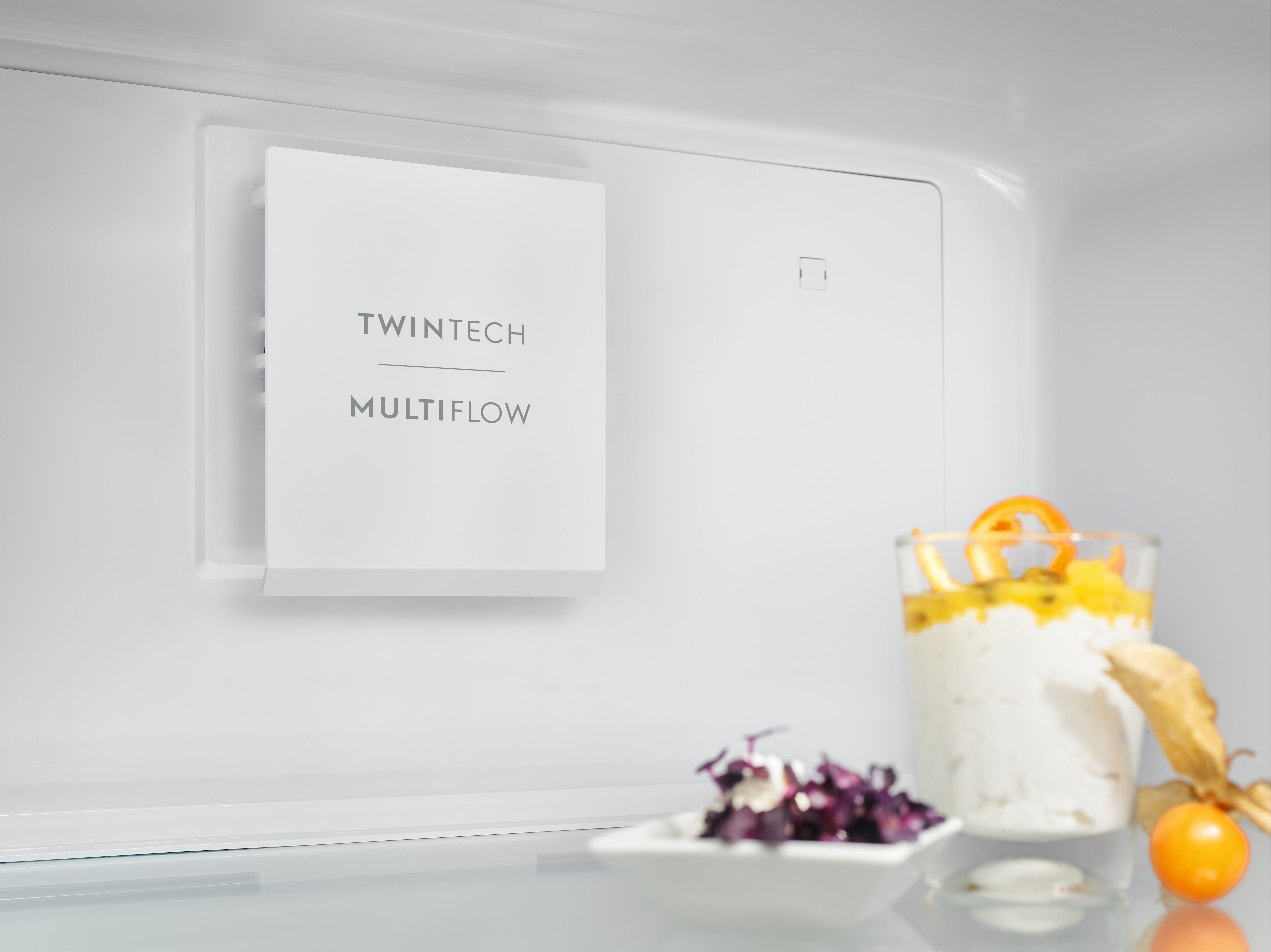 Beyaz bir buzdolabının içinde, bir TwinTech MultiFlow paneli ve bir bardakta tatlı görülüyor.