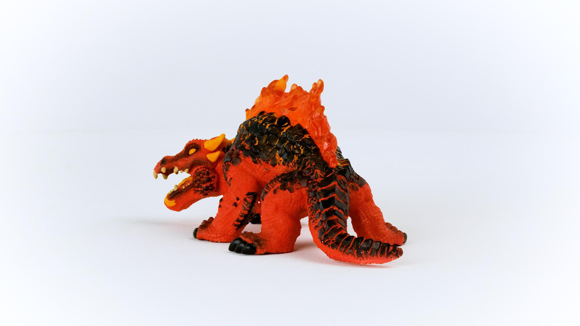 Ein Spielzeugdrache mit rotem und orangefarbenem Farbschema. Er steht auf einer weißen Oberfläche. Der Drache hat Hörner und Zähne.