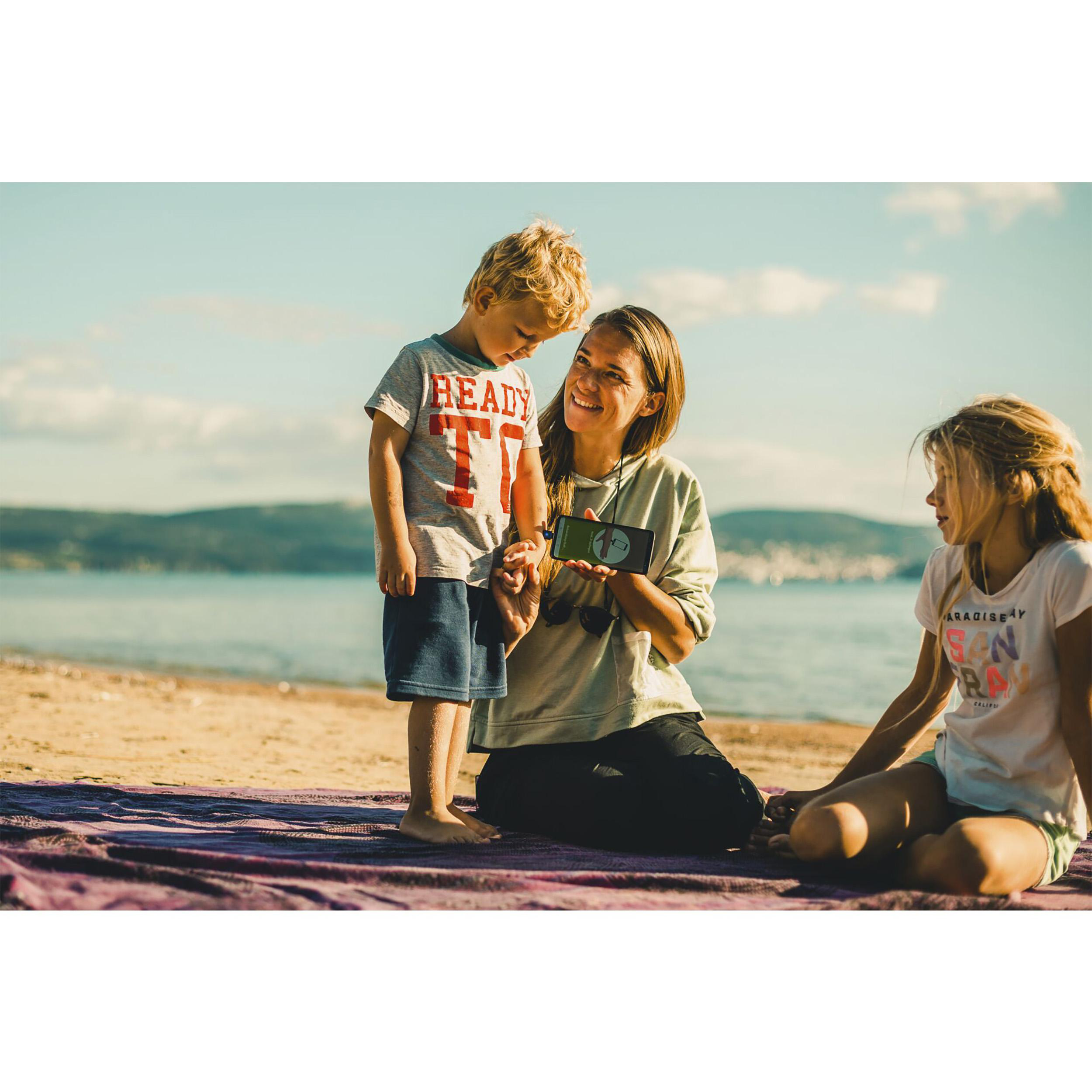 Eine Frau mit zwei Kindern am Strand, die auf ein Telefon schauen. Sonniger Tag, blauer Himmel und Meer im Hintergrund.