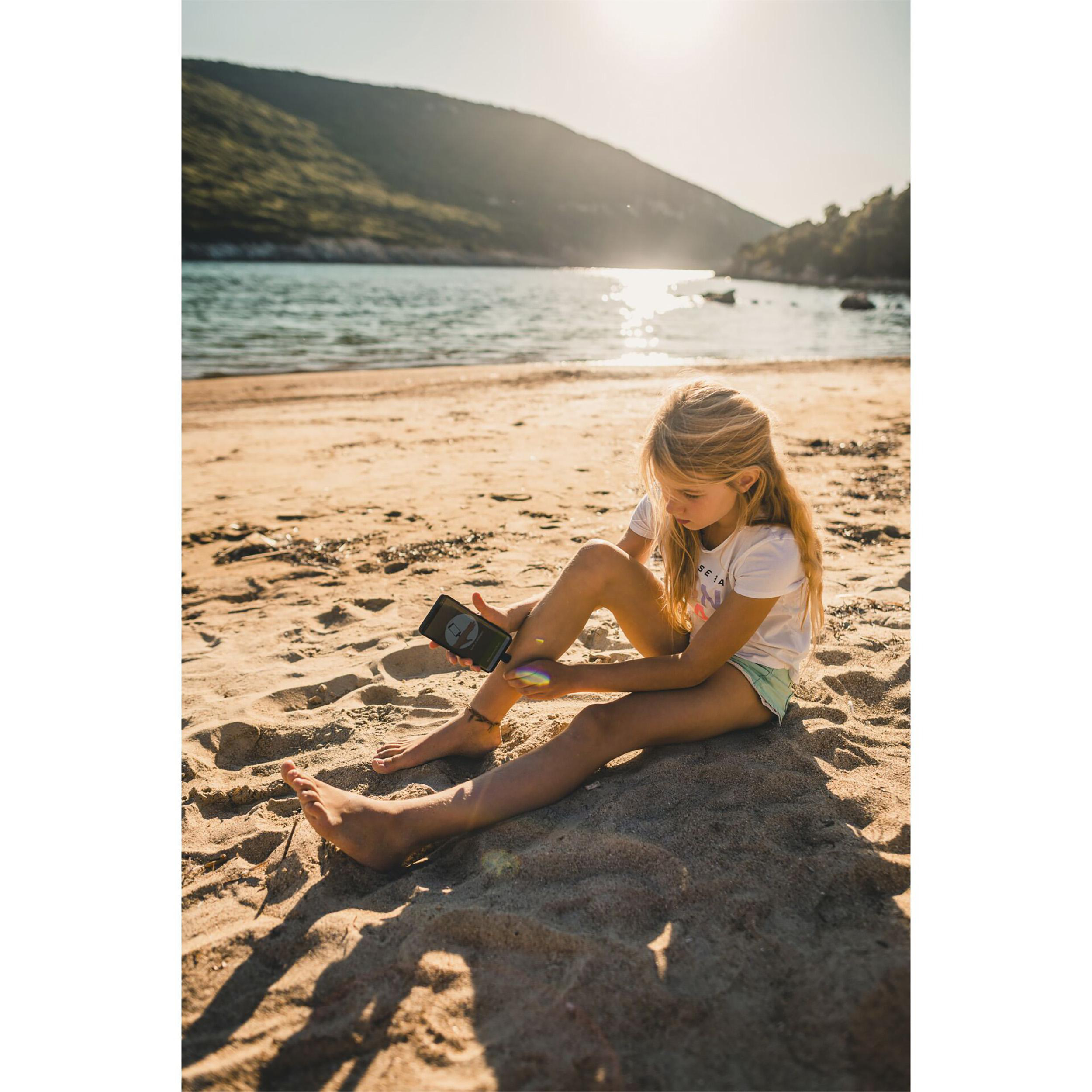 Ein Mädchen sitzt am Strand und schaut auf ihr Handy. Die Sonne geht unter.