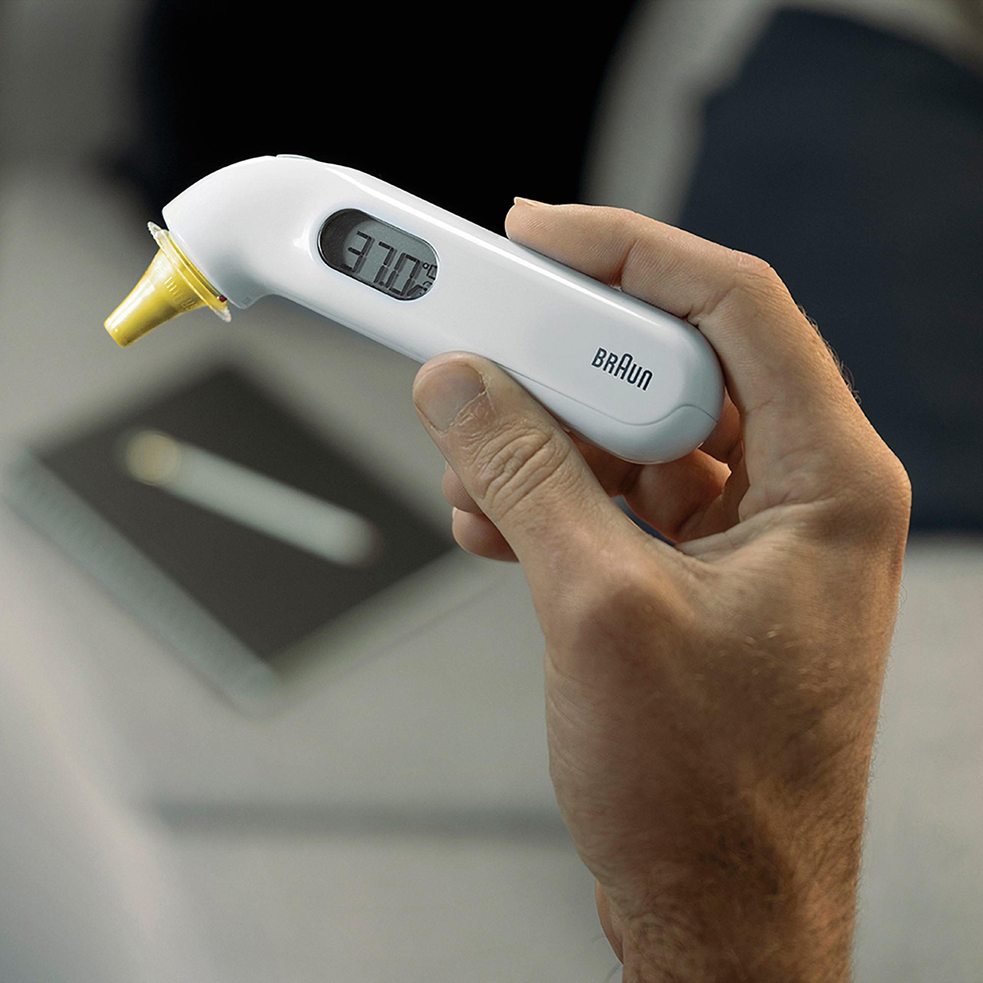 Hand hält weißes Braun-Ohrthermometer, Anzeige zeigt 37,0°.