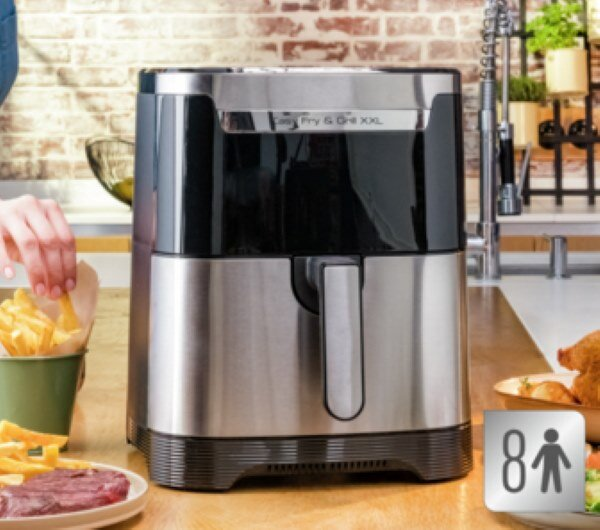 Frytownica powietrzna na blacie. Ręka wkłada frytki do zielonej miski. Stek z boku.