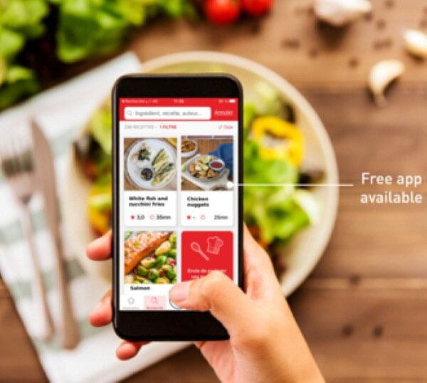 Ręka trzyma telefon z aplikacją kulinarną z przepisami, obok talerza z jedzeniem.