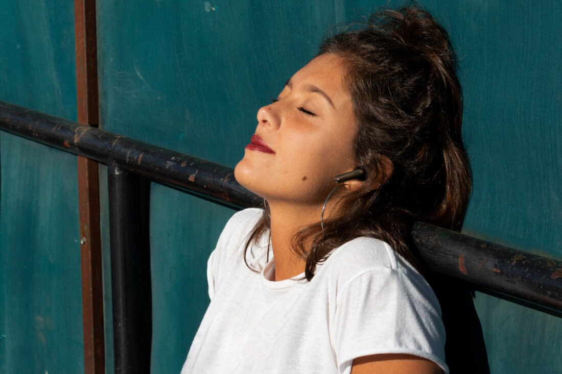 Kobieta z zamkniętymi oczami, w białej koszuli i słuchawkach, oparta do tyłu.